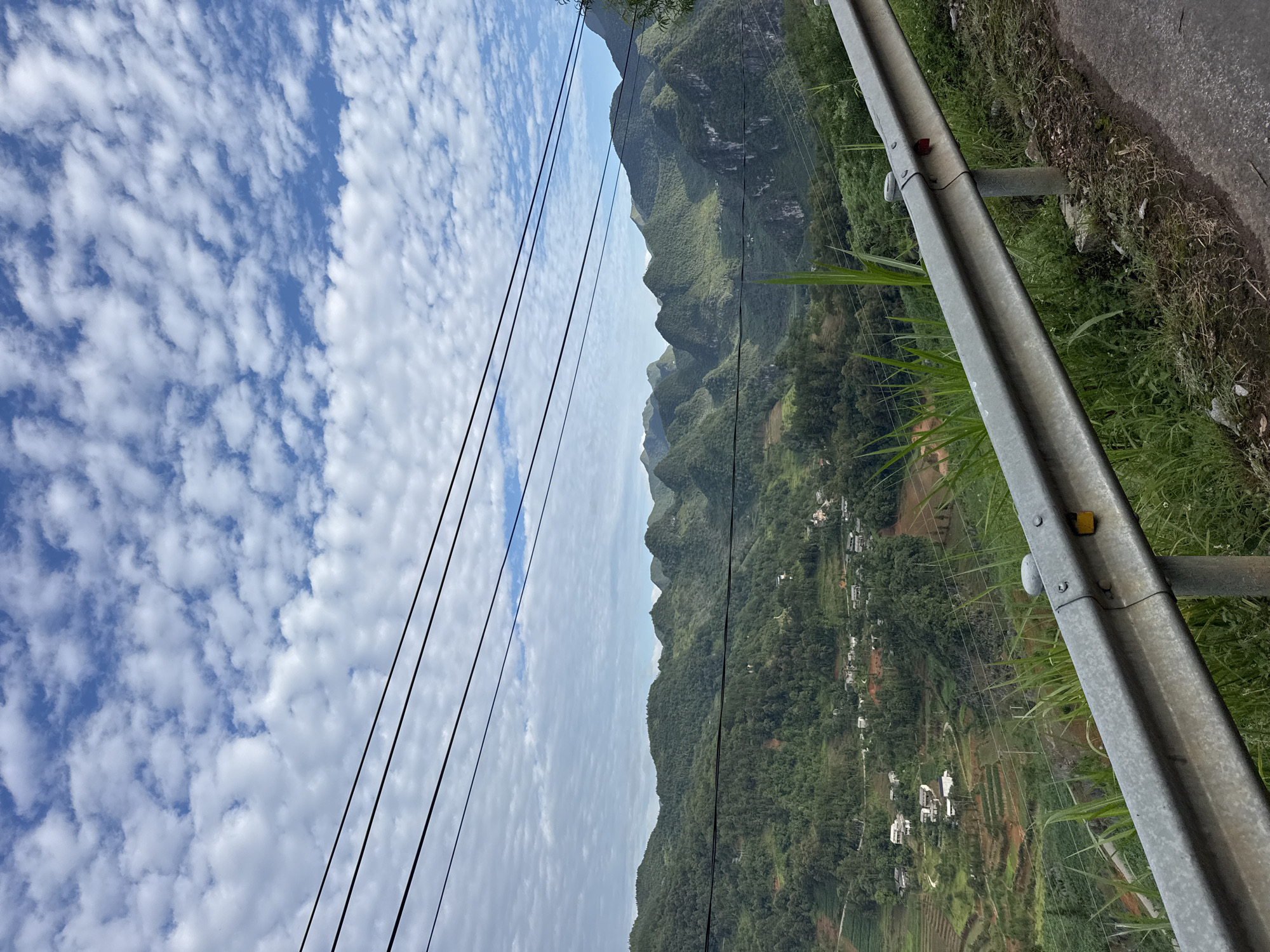 The image shows a landscape in Lũng Cú Commune, Vietnam. The sky is partially cloudy with a mix of blue and white clouds. A series of mountains extends across the middle of the image. Power lines are visible in the sky. A metal guardrail runs along the right side of the frame, positioned on the edge of a road. Green vegetation is growing around the guardrail. Below the mountains, there is a village or town with buildings and terraced fields.