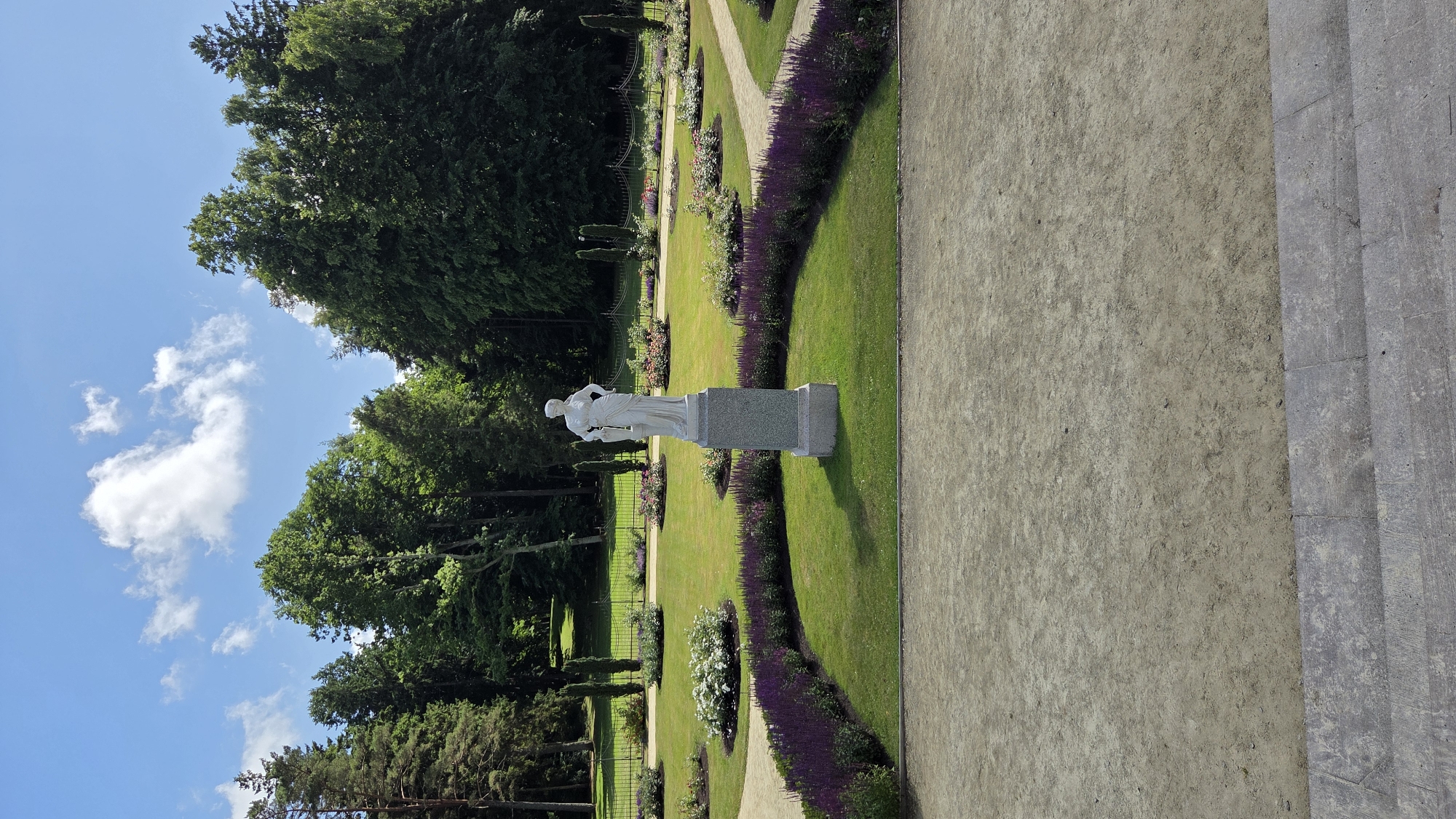 The image depicts a sunlit day in a meticulously maintained park, likely in Palanga, Lithuania, given the context of the original prompt. In the foreground, a wide, light-colored gravel path leads up to a stone staircase, suggesting a viewing platform or elevated area. Dominating the mid-ground is a classical white marble statue of a female figure standing on a grey stone pedestal. The statue is positioned centrally within a circular arrangement of vibrant purple flowers, bordered by a well-manicured green lawn. Beyond this, a series of curved pathways wind through the park, lined with more flowerbeds of various colors, including pink roses and white hydrangeas. Tall, dark green cypress trees are strategically placed throughout the landscape, adding verticality and structure. In the background, a dense, verdant forest forms a natural backdrop, with mature deciduous and coniferous trees. A dark metal fence runs along the edge of the park, separating it from the forest. The sky is a bright blue with scattered white clouds, indicating fair weather. The overall scene conveys a sense of tranquility and order within a beautiful natural setting.