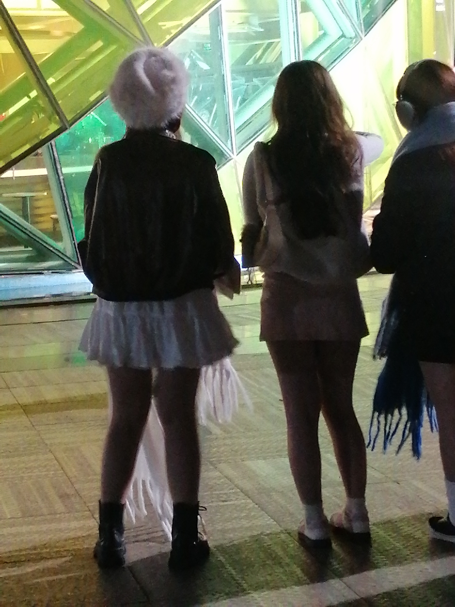 The image depicts three individuals standing on a tiled floor in front of a faceted, brightly colored structure, possibly a building facade or art installation. The light filtering through the structure casts a green and yellow glow on the surroundings.
The person on the left is viewed from the back, wearing a dark jacket over a white fluffy hat and a white pleated skirt. They are also wearing black boots.
In the center, another individual is seen from the back, with long, dark hair, wearing a light-colored top and a dark skirt. They appear to be holding a white bag.
The person on the right is also seen from the back, wearing dark clothing and a blue scarf. They have headphones on their ears and are wearing white socks with dark shoes.
The setting suggests an outdoor or semi-outdoor urban environment. There are no explicit visual cues to pinpoint the location within Da Lat, Vietnam, such as specific landmarks, signage, or architectural styles that are uniquely identifiable to Da Lat. The overall impression is of a contemporary, potentially artistic or commercial urban space.