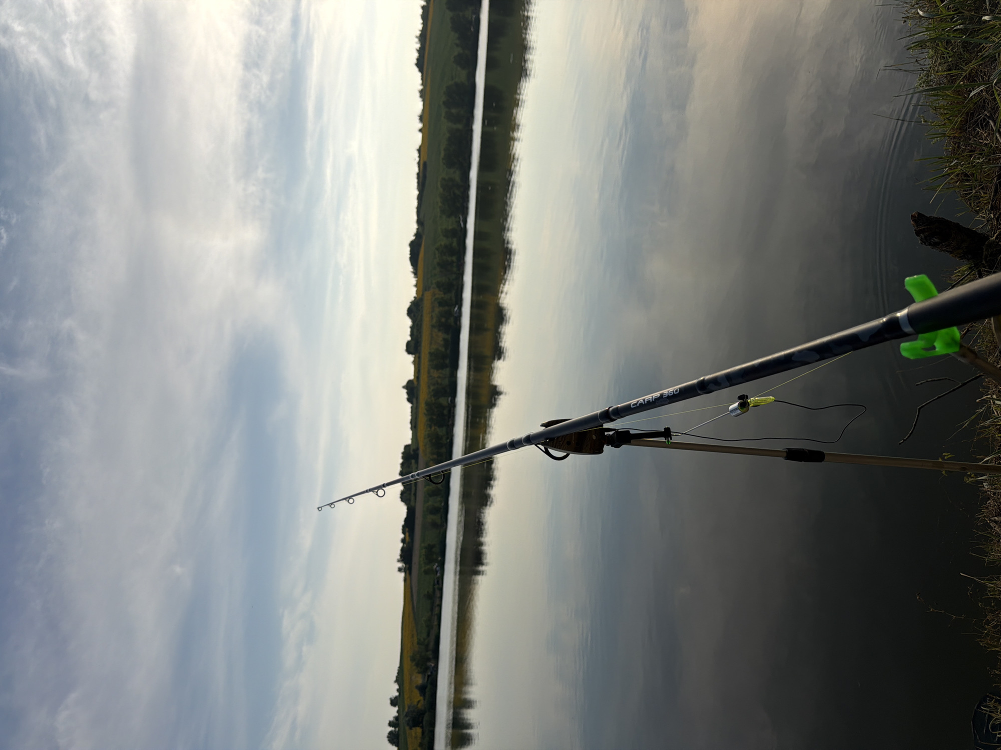A fishing rod is propped up on a tripod by the edge of a calm lake. The rod is angled towards the water, with a fishing line extending into the distance. The lake's surface reflects the sky, which is filled with wispy clouds. In the background, a lush green treeline borders the water, and beyond that, rolling hills are dotted with yellow flowers, likely sunflowers. The overall scene suggests a peaceful and serene environment, possibly in the countryside near Găleștii Noi, Moldova, given the agricultural landscape. There are no people visible in the image.