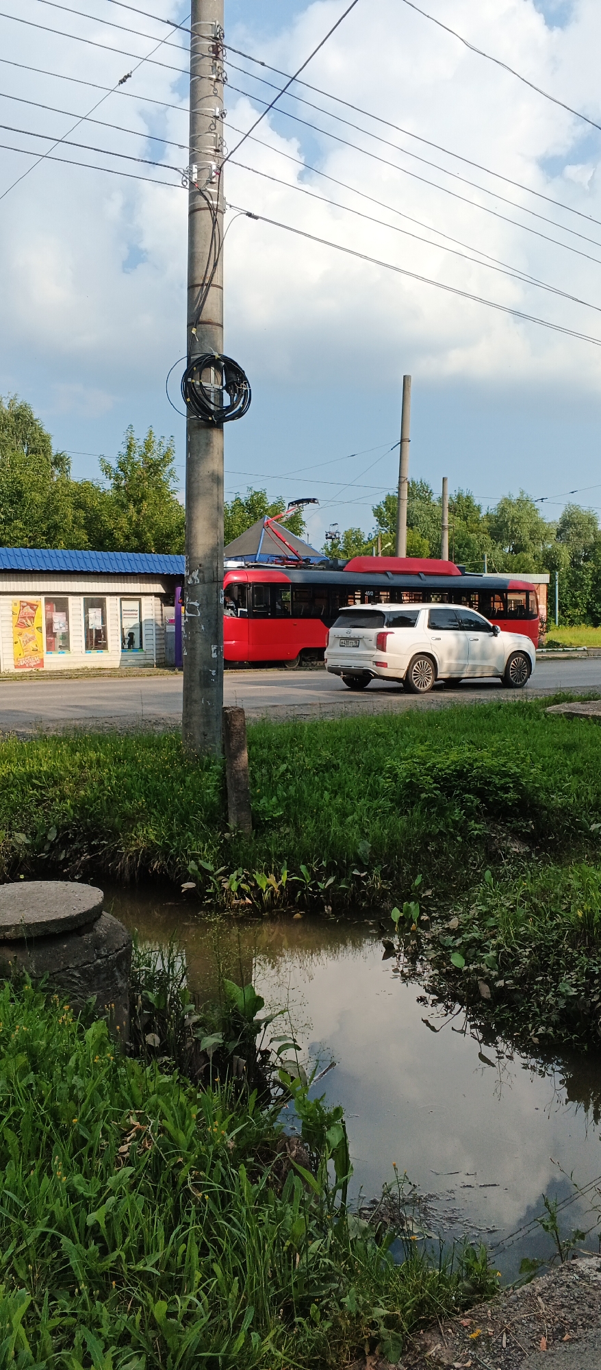 This image captures a street scene in Izhevsk, Russia, likely on a sunny day with scattered clouds. In the foreground, lush green grass grows alongside a narrow, shallow body of water, possibly a drainage ditch, with reflections of the sky and surrounding vegetation visible on its surface. A concrete manhole cover is partially submerged in the grass to the left.
Dominating the middle ground is a concrete utility pole, a common sight in urban environments, from which numerous electrical wires extend across the frame. A coiled bundle of black cable is attached to the pole. To the left of the pole is a small, single-story structure with a blue corrugated metal roof. The building appears to be a kiosk or small shop, with windows displaying advertisements and goods.
Further back, a bright red and dark blue tram travels along a track. The tram has its pantograph extended, indicating it is drawing power from the overhead lines. To the right of the tram, a white SUV is parked on the side of the road, facing away from the viewer. The vehicle's license plate is partially visible. The background consists of a line of mature green trees, suggesting a suburban or a less densely built-up urban area. The overall impression is one of everyday life in a Russian city, with elements of public transportation, commercial activity, and natural elements coexisting.