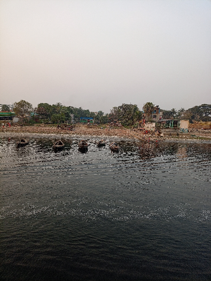 The image portrays a heavily polluted river scene in what appears to be an urban or semi-urban area of Zazira, Bangladesh. The foreground is dominated by dark, murky river water, characterized by numerous ripples and patches of white foam or bubbles, indicative of poor water quality. Approximately six small, dark boats, likely wooden, are visible on the water, some appearing partially submerged or laden with items.
The riverbank is extensively covered with a thick layer of accumulated trash, debris, and refuse, stretching from the waterline to the base of the structures and trees further inland. Beyond the littered bank, a mix of sparse green trees and simple, somewhat dilapidated buildings can be seen, with some structures exhibiting colorful elements like blue and green. Several small, indistinct figures, possibly people, are discernible on the riverbank near the buildings, though their specific activities are not clear.
The sky is a uniform, pale, hazy grey, suggesting either early morning, late afternoon, or an overcast/smoggy day, with diffused lighting casting no strong shadows. The scene conveys a strong impression of significant environmental degradation and waste management challenges. No visible text is present.