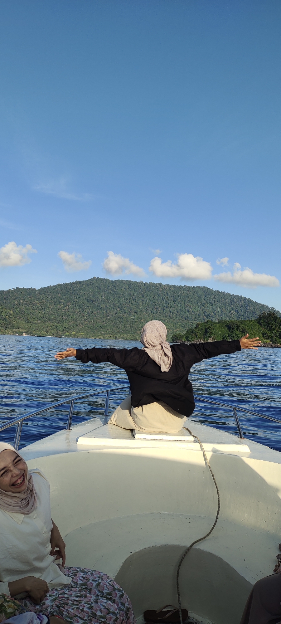 In Sabang, Indonesia, two women are on a boat in the ocean. One woman, wearing a black jacket and beige pants with a headscarf, sits facing away from the camera with her arms outstretched, seemingly embracing the view. The other woman, in the foreground, is wearing a white shirt and a headscarf, and she is laughing. The boat is white and is on the water, which is blue and wavy. In the background, a lush green mountain is visible under a bright blue sky with scattered white clouds.