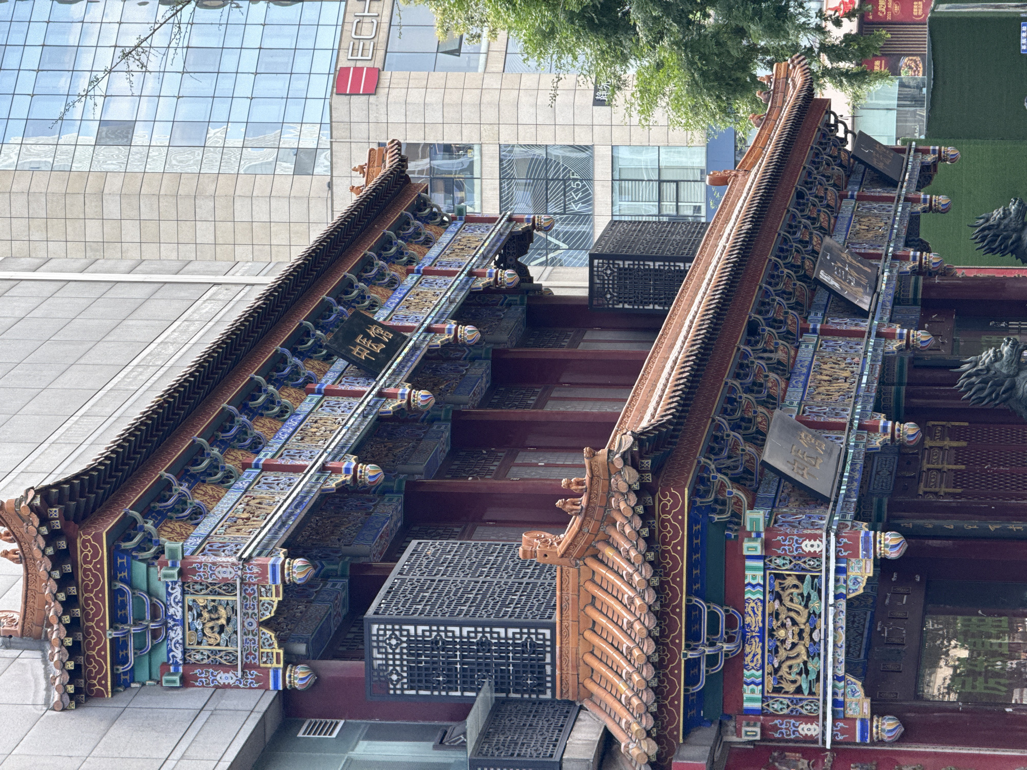 A ground-level, medium-shot photograph depicts traditional Chinese architecture juxtaposed with a modern office building, taken in the Jinjiang District of China. The traditional structure exhibits a tiered roof with detailed decorative elements in red, blue, and gold. Intricate latticework boxes are attached to the building. To the left, a section of the structure is adjacent to a plain gray surface. Behind the traditional building, a contemporary office building with glass windows rises, reflecting the sky and surrounding structures. The photograph is taken from a perspective where the traditional architecture is in the foreground and slightly tilted, creating a dynamic composition with the modern building in the background.