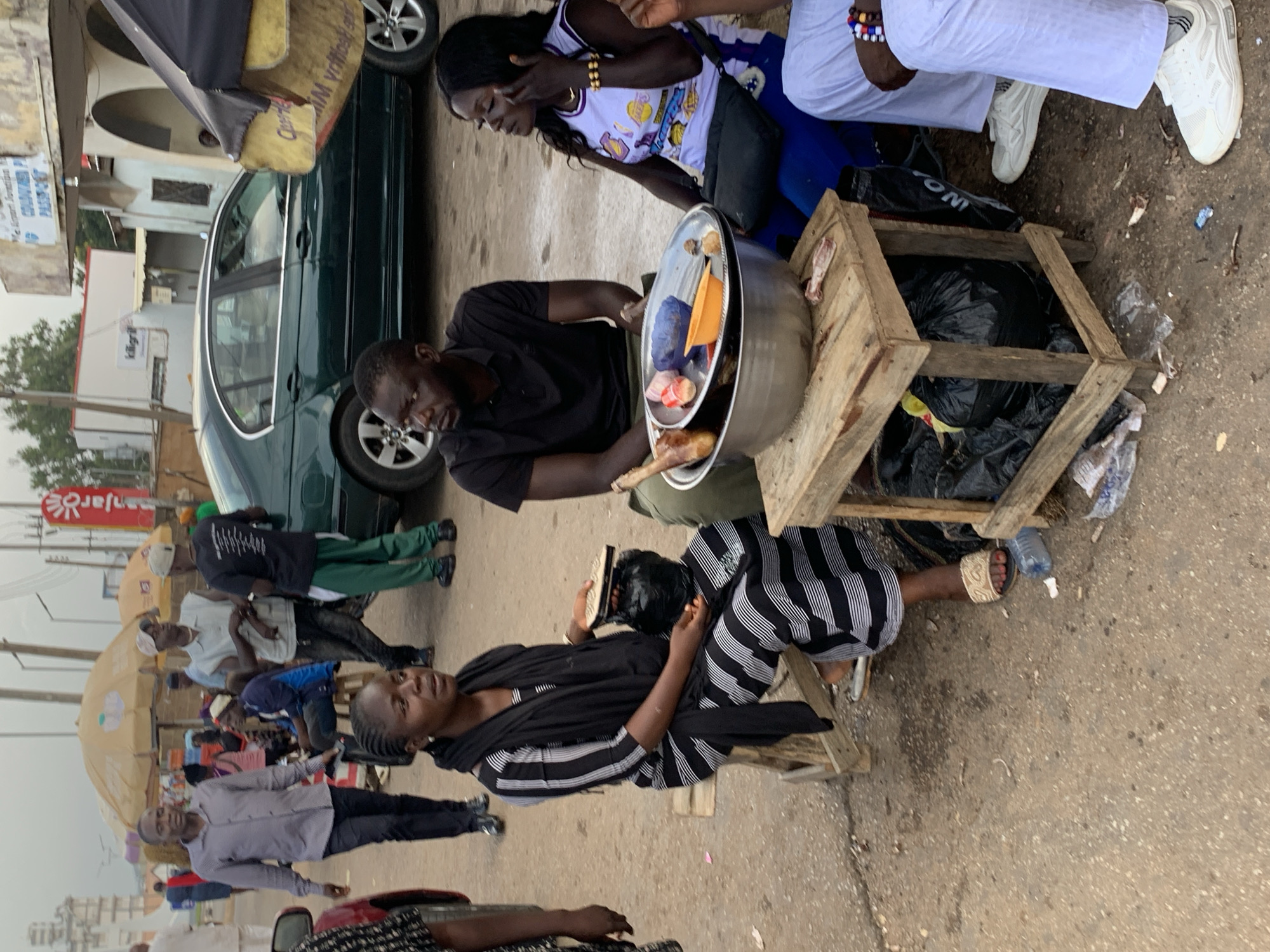 In an outdoor scene in Jos, Nigeria, several individuals are gathered near a roadside food stall. A woman in a black and white striped dress and headscarf sits on a wooden stool, holding a small container and a plastic bag. Beside her, a man in a black polo shirt is seated, looking to the side. Another woman in a white and purple tank top is seated with her hand touching her face, and a man in white clothing is also seated to her right. In the foreground, a metal bowl filled with food items, including what appears to be meat and vegetables, sits on a small wooden table laden with black plastic bags. A green sedan is parked in the background, and other people and market stalls are visible further down the street, suggesting a bustling urban environment. The ground is paved with asphalt and shows signs of wear and tear, with scattered debris.