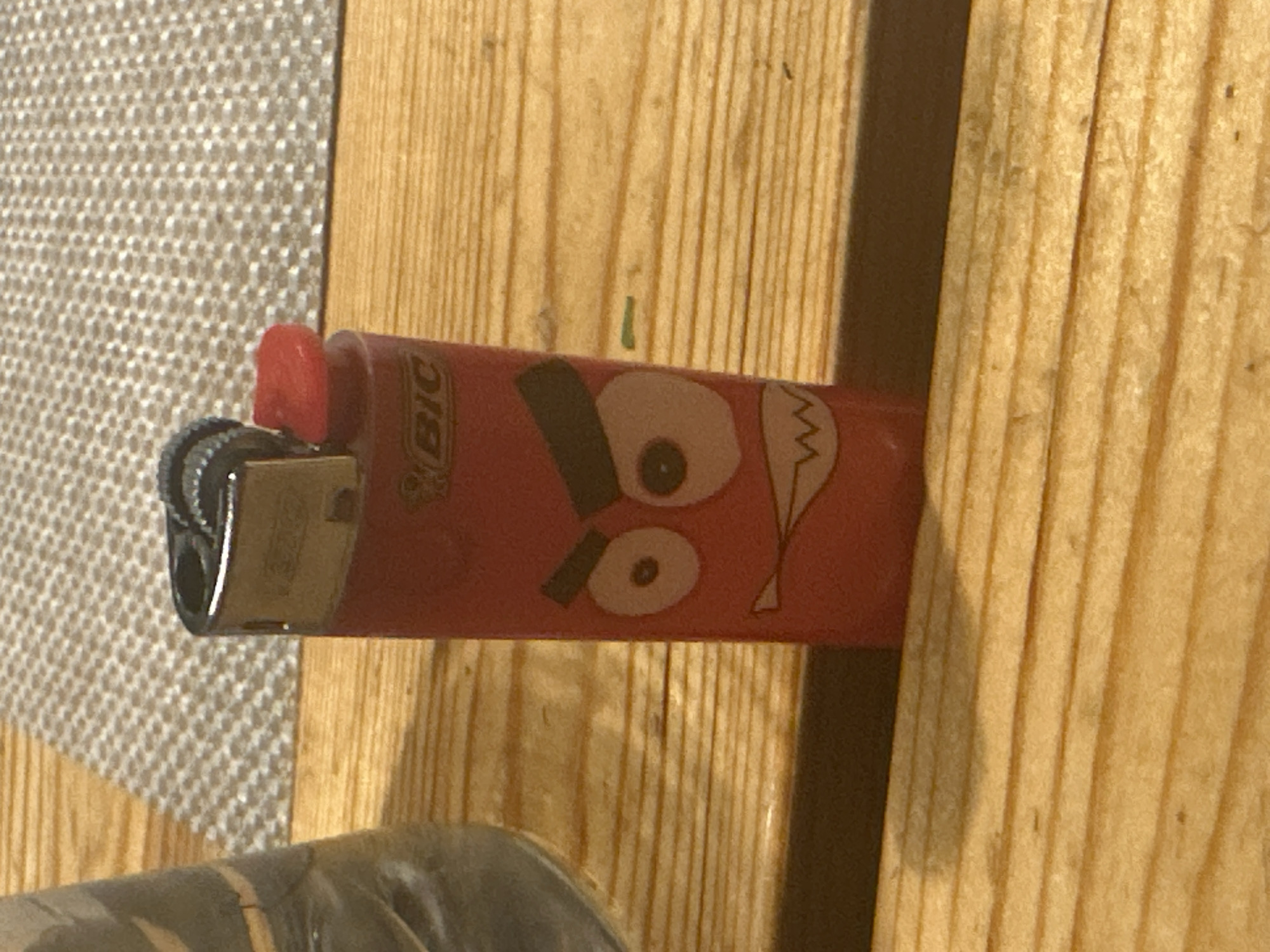 A close-up shot features a red BIC lighter with a cartoonish angry face printed on it, standing upright on a wooden surface. The lighter has a metallic top with a fluted wheel and a red ignition button. The face on the lighter consists of two large, wide-eyed circles with black pupils, and angry, slanted eyebrows. Below the eyes, a jagged, W-shaped mouth with sharp teeth is depicted. The background shows a portion of a glass object on the left and a textured surface, possibly a placemat, behind the lighter. The wooden surface appears to be a table with visible grain and some subtle marks. The lighting suggests an indoor setting, likely with artificial light casting a soft shadow to the right of the lighter. There are no people or animals present in the image. The scene is static and devoid of any activity. The location in Востец Снт, Russia is not determinable from the visual information provided.