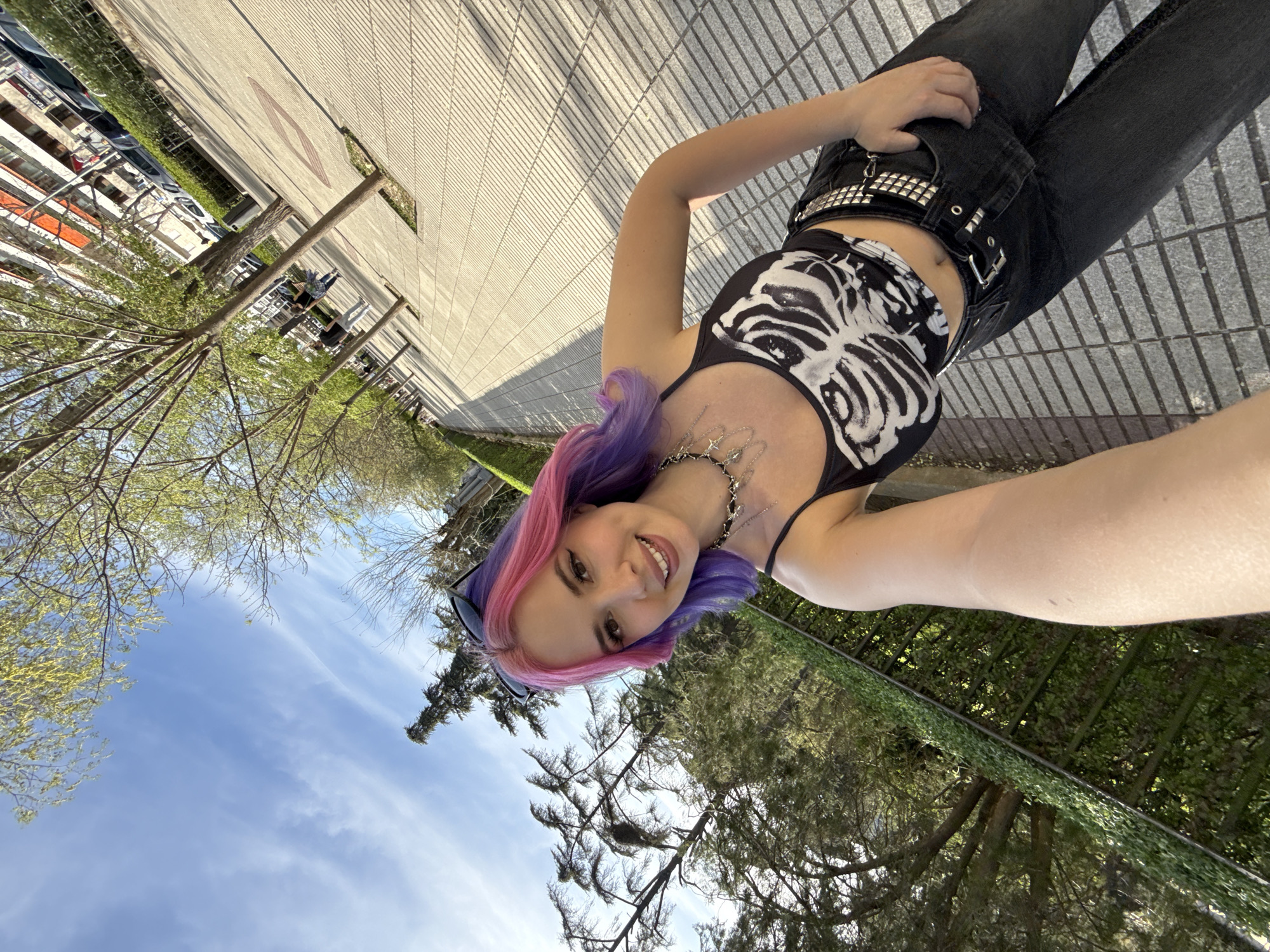 A young woman with pink and purple hair is standing on a paved surface, likely a sidewalk or plaza, and taking a selfie. She is wearing a black skeleton-print tank top, black jeans, and a studded belt. A silver necklace adorns her neck. In the background, there is a row of trees and a building. The sky is visible in the upper left corner. Based on visual landmarks and architectural styles, the location may be the city of Madrid, Spain.