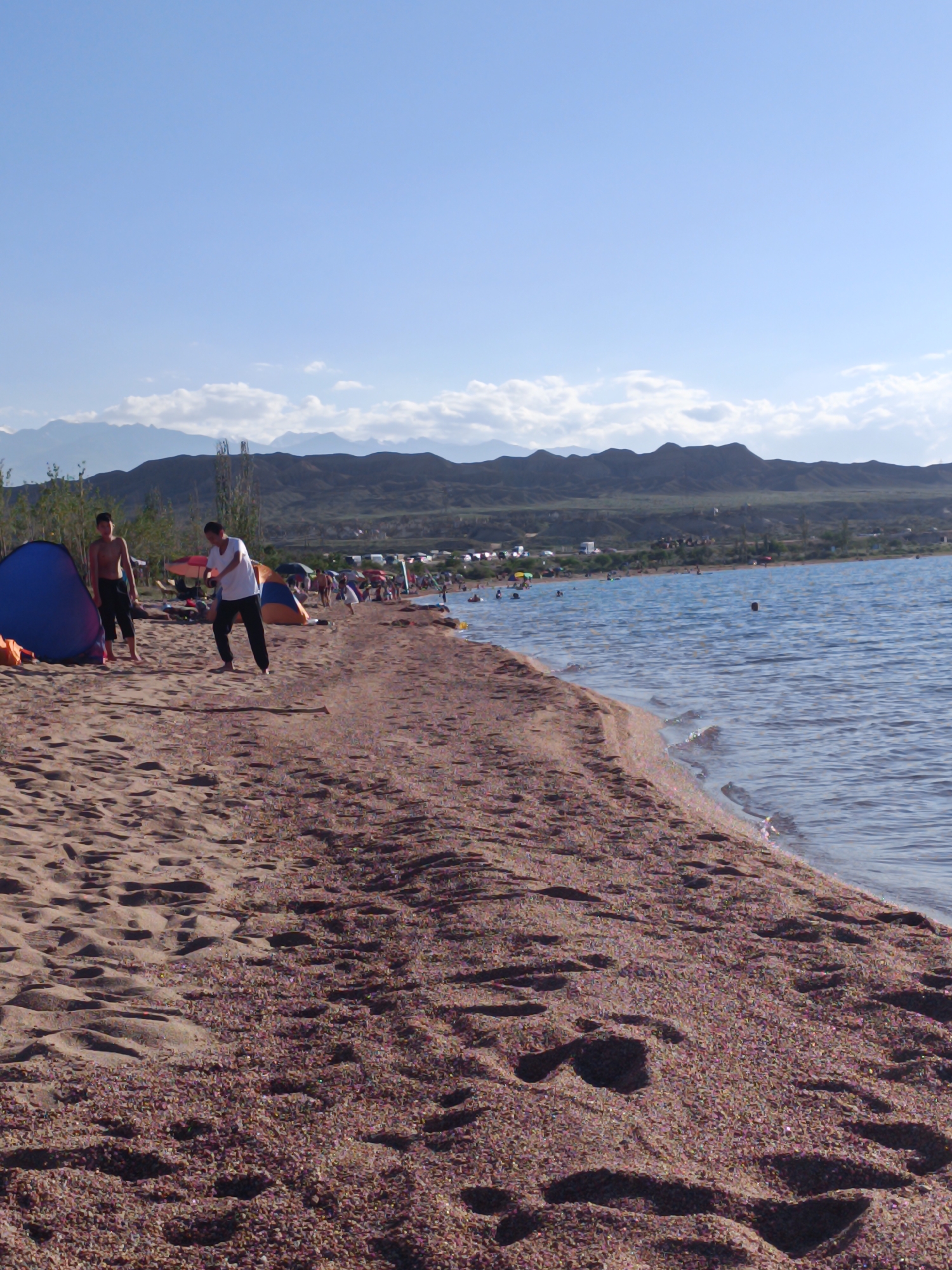 The image depicts a sunny day on a pebble and sand beach along a large body of water, likely Lake Issyk-Kul, near Tong, Kyrgyzstan. In the foreground, two young men are on the beach. One, with a darker complexion and dark hair, is standing near a blue tent, wearing dark shorts. The other, with lighter skin and dark hair, is wearing a white t-shirt and dark pants, and appears to be in motion, possibly playing or demonstrating something. Behind them, the beach is lined with various tents and umbrellas, suggesting a recreational area. Further in the background, many people are visible, some in the water, which is a clear blue. The shoreline recedes into the distance, with mountains forming the backdrop. The sky is clear and blue, with some wispy clouds. The environment is characterized by the sandy beach, the calm water, and the mountainous landscape.