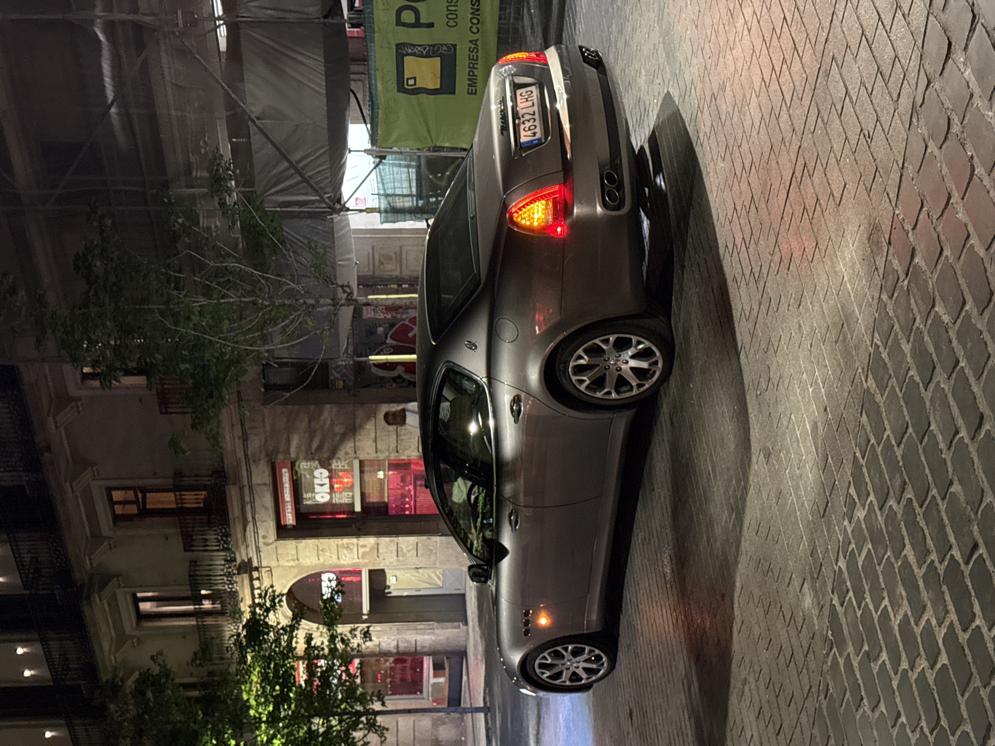 The image depicts a silver-grey Maserati GranTurismo parked on a cobblestone street in Barcelona, Spain, at night. The rear of the vehicle is visible, showcasing a license plate reading "4632 LHG". The car is parked in front of a building with illuminated signage. One sign says "OKIO". Scaffolding covered in translucent sheeting is visible in the background. A tree with green foliage partially obscures the building.