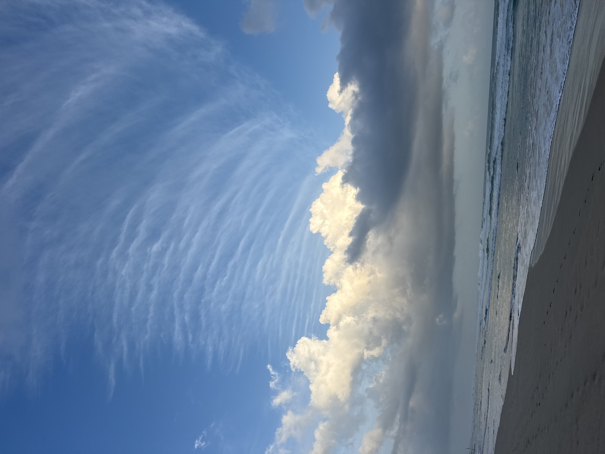 This is a landscape image of a beach with a dramatic sky. The scene is dominated by the sky, which takes up about two-thirds of the frame. On the left side, a vast expanse of clear blue sky is filled with wispy, streaky clouds that form a delicate, fan-like pattern. These clouds are thin and white, suggesting high altitude.
As the eye moves to the right, the sky becomes more dramatic with larger, denser clouds. A towering formation of cumulus clouds, with bright white tops and darker, shadowed undersides, juts into the frame from the right. These clouds appear to be illuminated from the side, possibly by the sun. Further to the right, the sky is overcast with dark, gray clouds, suggesting an approaching weather system or the lingering effects of one.
The bottom third of the image shows a beach and the ocean. The ocean is calm, with gentle waves rolling onto the shore. The water appears to be a muted gray-blue, reflecting the overcast sky. The beach is a strip of sand, light brown in color, with a few faint footprints visible. The horizon line is not straight, giving the image a slightly tilted appearance.
The time of day appears to be late afternoon or early evening, given the lighting on the clouds and the overall mood of the scene. The weather is mixed, with patches of clear sky and areas of heavy cloud cover. The location appears to be a tropical beach, with the presence of the ocean and sand. There are no people or man-made objects visible in the frame. The overall emotion evoked by the image is one of awe and natural beauty, with a sense of impending change suggested by the contrasting cloud formations.
