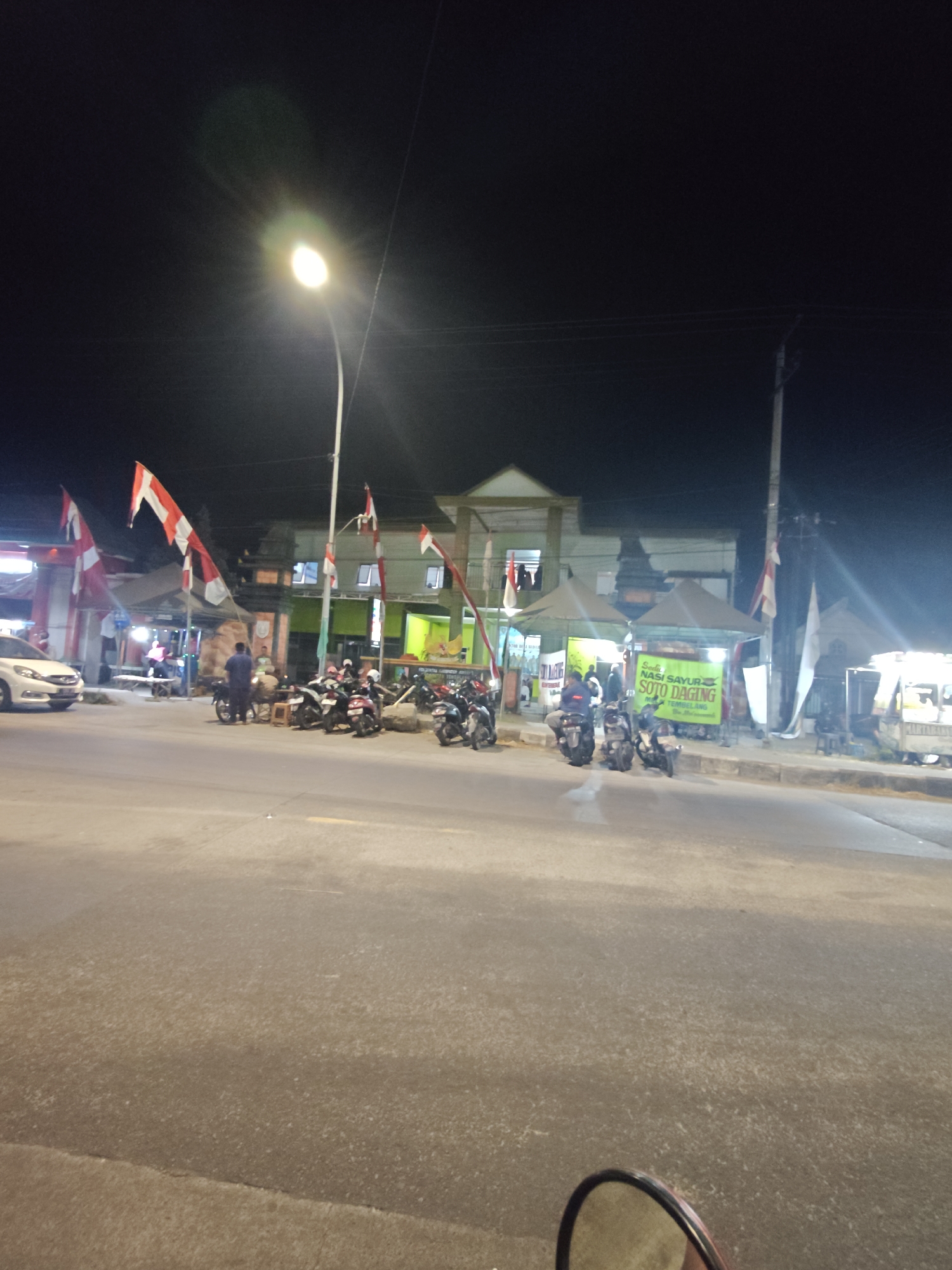 At night, a street scene unfolds in Bedah Lawak, Indonesia. A white car is parked on the left side of the road. Several motorcycles are parked in a row. Flags, red and white, are displayed prominently on poles. Buildings line the street, some with awnings providing shelter. A street lamp illuminates the scene. People are visible standing and interacting. The road surface is asphalt.