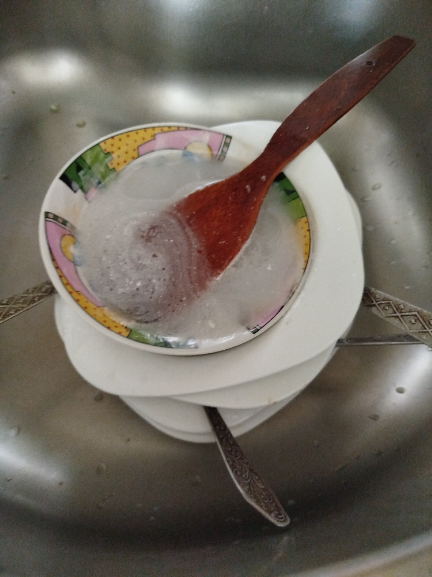 In a stainless steel sink, a stack of white plates is partially submerged in soapy water. A wooden spoon rests on top of a small, decorative bowl, which is also filled with soapy water and placed on the stack of plates. A patterned metal spoon lies at the bottom of the sink. The scene appears to be a domestic setting, likely a kitchen sink in a home or public facility. The location is identified as Kryvaselski Sel'ski Sovet, Belarus.