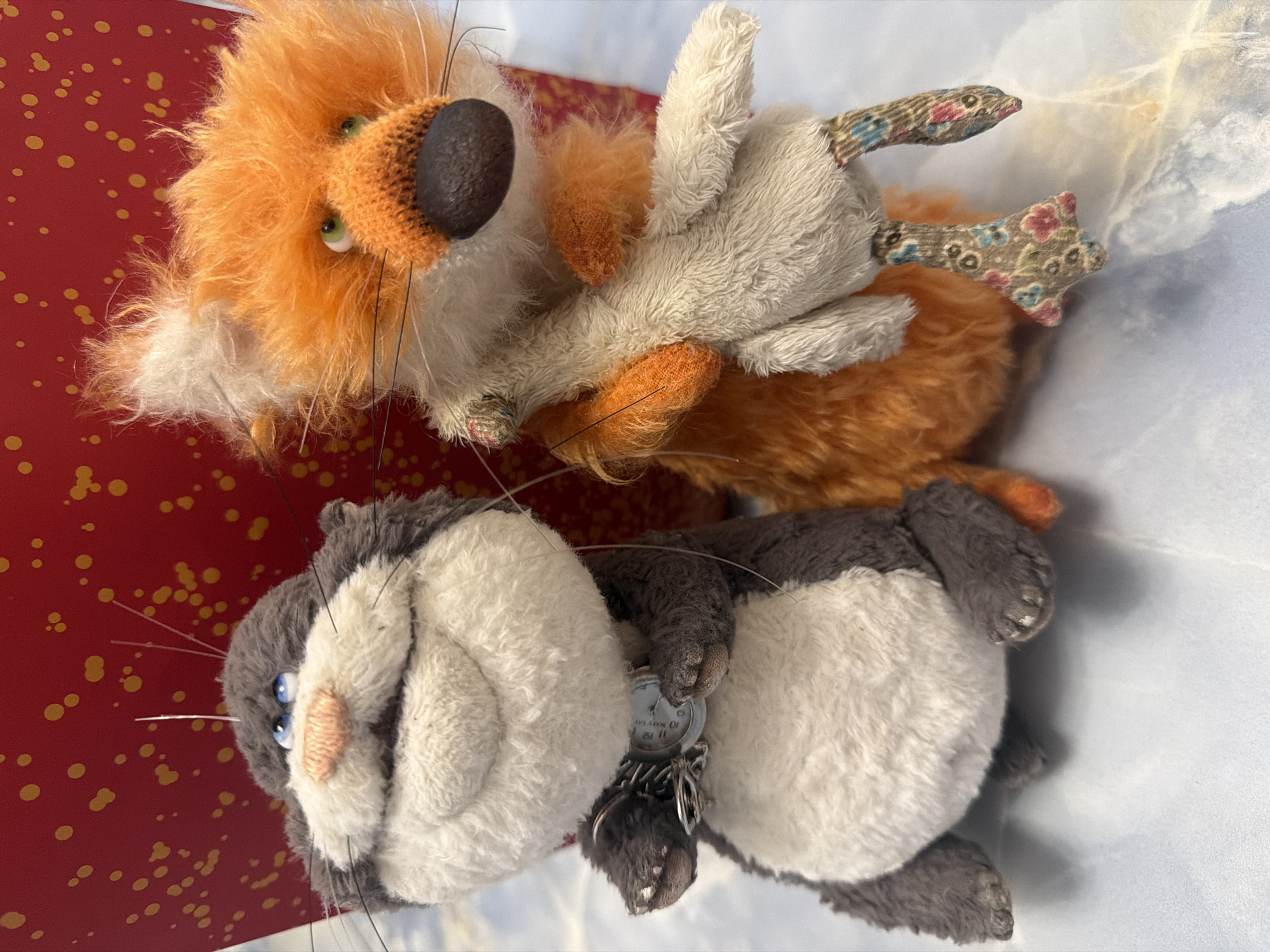 Two stuffed animal toys are positioned on a surface. On the left is a gray and white cat-like toy with blue eyes, a pink nose, and whiskers. It wears a silver-colored chain with a circular pendant around its neck. To the right is an orange fox-like toy with green eyes and a dark nose. It is holding a beige stuffed bird with its paws. The background features a red surface with yellow speckles.