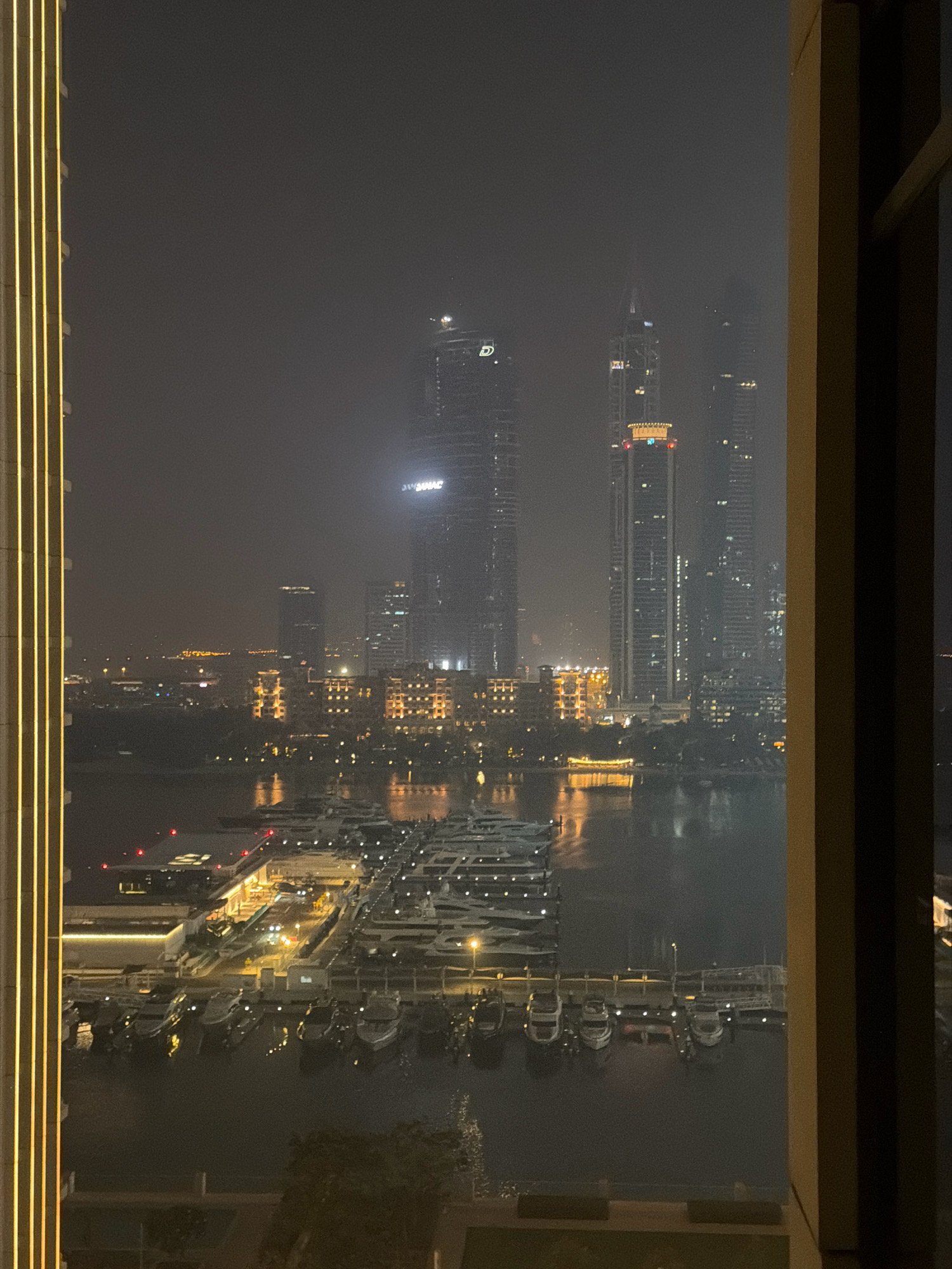 This is a night-time, vertical, full-shot photograph taken from an elevated perspective, looking out of a window frame with visible vertical trim on the left and right. The scene depicts a marina in Dubai, United Arab Emirates, characterized by a dark, misty atmosphere with numerous illuminated skyscrapers and a body of water filled with moored yachts and boats.
The foreground features the window frame, with vertical golden light strips on the left and a broader, darker frame on the right. Beyond the window, a wide expanse of water, likely a marina, dominates the lower half of the image. Moored yachts and smaller boats are densely packed in the water, their lights reflecting on the surface. Some of these vessels appear to be larger yachts with multiple decks, while others are smaller recreational boats.
In the middle ground, a modern marina structure is visible with illuminated pathways and buildings. Red lights are present on some structures in the marina, possibly indicating navigation markers or safety lights. Beyond the marina, the city skyline is prominent. Tall, slender skyscrapers pierce the misty night sky. Many of these buildings are illuminated with lights visible through their windows, creating a sparkling effect. Some buildings have distinct architectural features and illuminated signage. One prominent skyscraper in the center-right has a golden illuminated top. Another skyscraper in the mid-distance has a horizontal illuminated section near its peak. The haze or fog in the atmosphere softens the details of the distant buildings, giving them a somewhat ethereal appearance.
The overall impression is of a vibrant, modern city at night, with a clear contrast between the illuminated man-made structures and the dark, possibly artificial, water body of the marina. The photographic quality suggests it was taken with a mobile device, as evidenced by some graininess in the image. No people are discernible in the photograph.