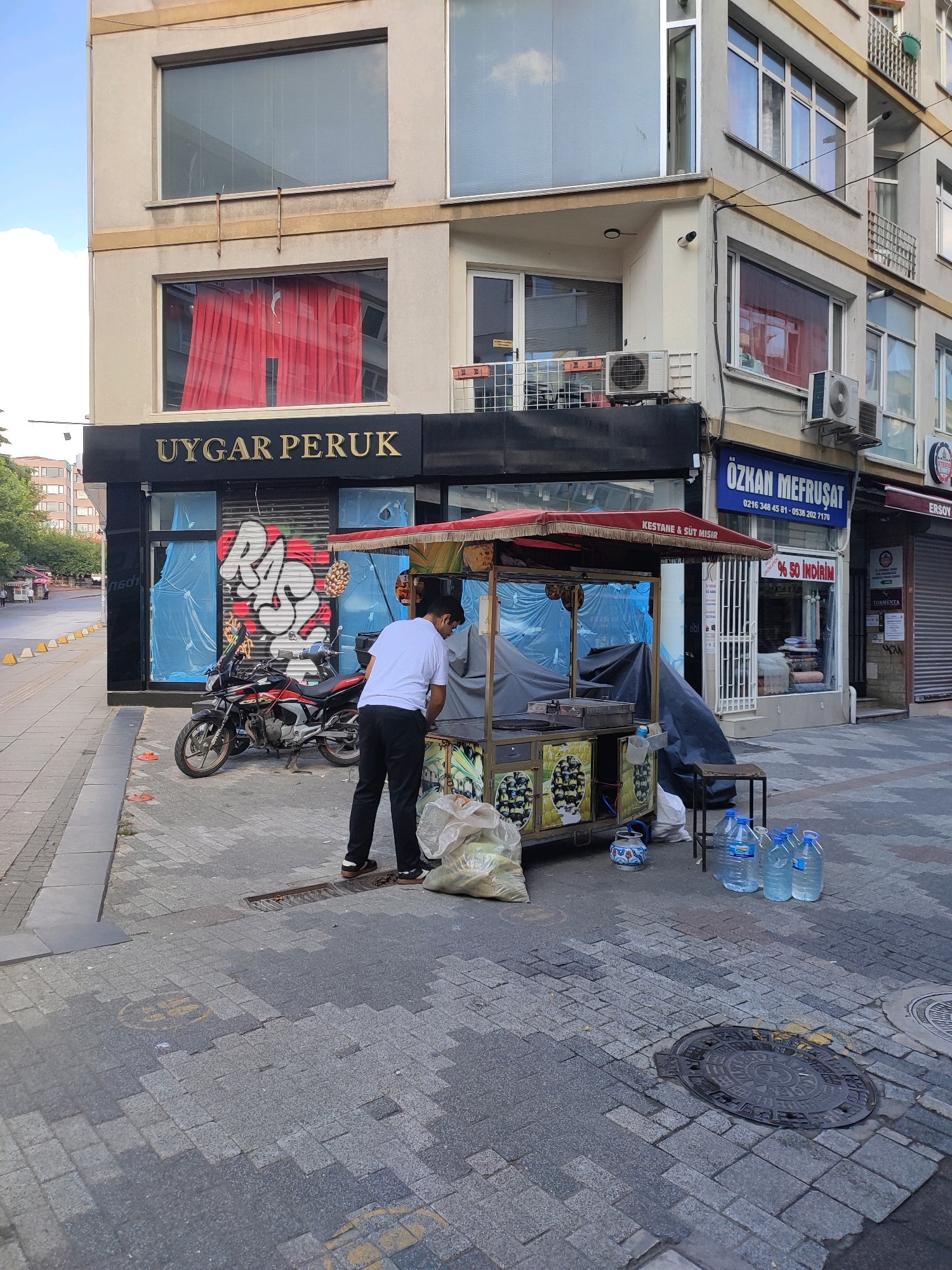 In Kadıköy, Turkey, a street vendor operates a cart selling roasted chestnuts and corn. The cart is positioned on a paved sidewalk in front of a commercial building. A person in a white t-shirt and black pants is attending to the cart. Behind the cart, a red motorcycle is parked, and the storefront windows are covered with blue plastic sheeting. The building above the storefront displays the name "UYGAR PERUK" in gold lettering. Across the street, other businesses are visible, including one with a blue sign that reads "ÖZKAN MEFRUŞAT" and a phone number. The street is lined with traffic cones, suggesting a pedestrian walkway. The overall scene depicts a typical urban commercial area.