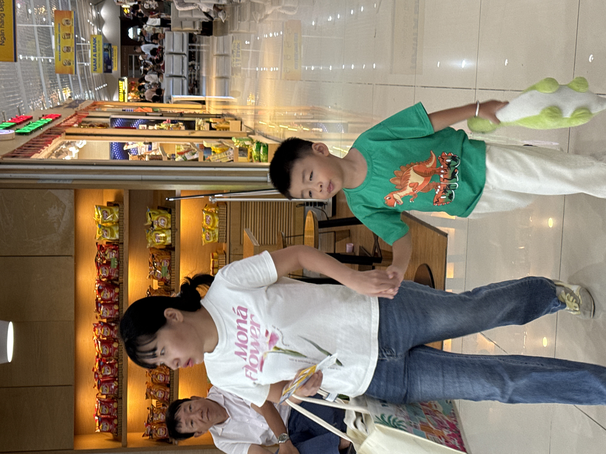 A woman and a young boy are walking through a brightly lit airport terminal, likely in Bac Cam Ranh Ward, Vietnam. The woman, wearing a white t-shirt with pink lettering and a floral graphic, and blue jeans, holds a shopping bag in her left hand and her right hand is interlocked with the boy's. The boy, dressed in a green t-shirt with a dinosaur graphic and white pants, is holding a stuffed dinosaur toy. They are walking away from the camera, towards the right side of the frame. In the background, shelves stocked with snacks are visible, along with other travelers and shops. The floor is a highly reflective surface, amplifying the overhead lighting.