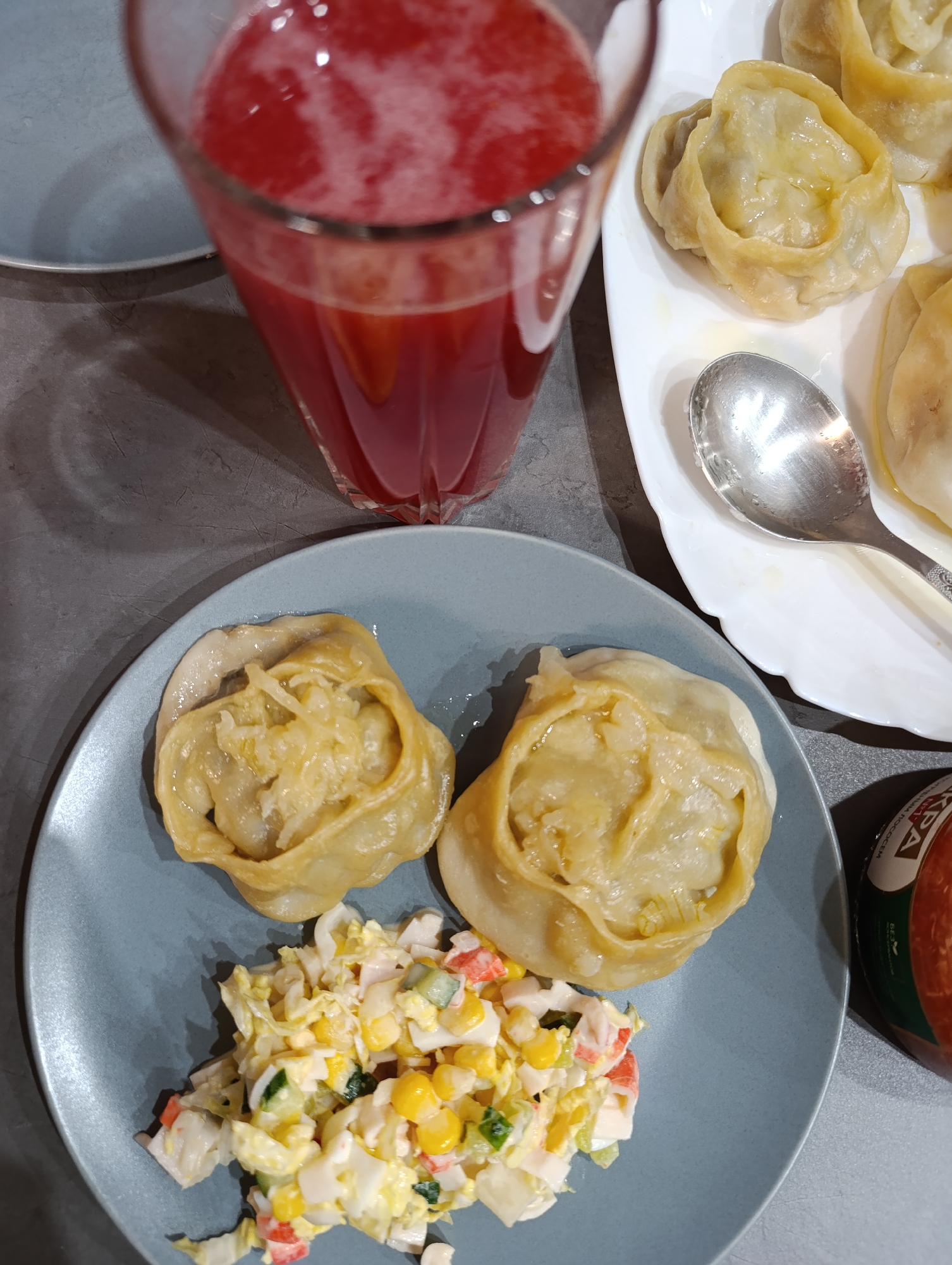 The image shows a table setting in what appears to be a residential setting. On a gray surface are two plates: a gray plate with two steamed dumplings (manti) and a mixed salad of cabbage, corn, cucumber, and crab sticks; and a white plate with several more manti, a spoon resting on it. There is also a tall glass of red liquid, likely juice or a compote. A jar with a red label, written in Russian, is visible on the right edge of the image. Given the Russian labeling on the jar, it is likely located in Russia. Although there is no information about the city of Chaikovsky, Russia in the image, I can't be certain that this location in Chaikovsky, Russia.