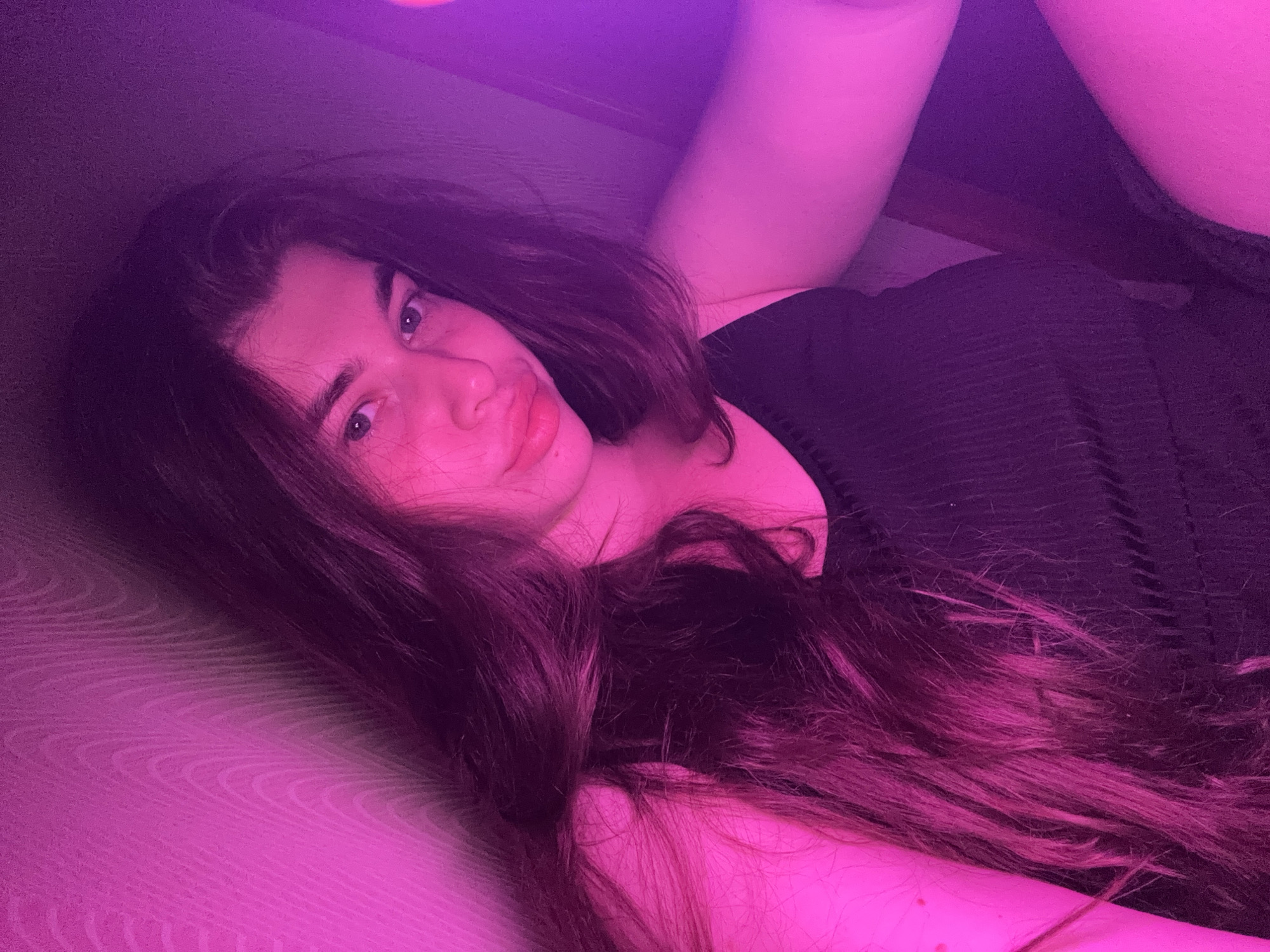 The image shows a young woman with long, dark hair, wearing a black tank top. She is leaning against a wall that has a textured surface. The scene is illuminated by pink and purple lighting, creating a dramatic and atmospheric effect. Her face is turned towards the camera, and she is looking directly at the viewer. The background is mostly out of focus, with the lighting creating a gradient of colors. There is no discernible location in the city of Мещёры, Russia, or any other specific location indicated in the image. The subject is alone and there are no other objects or people present. The woman appears to be taking a selfie.