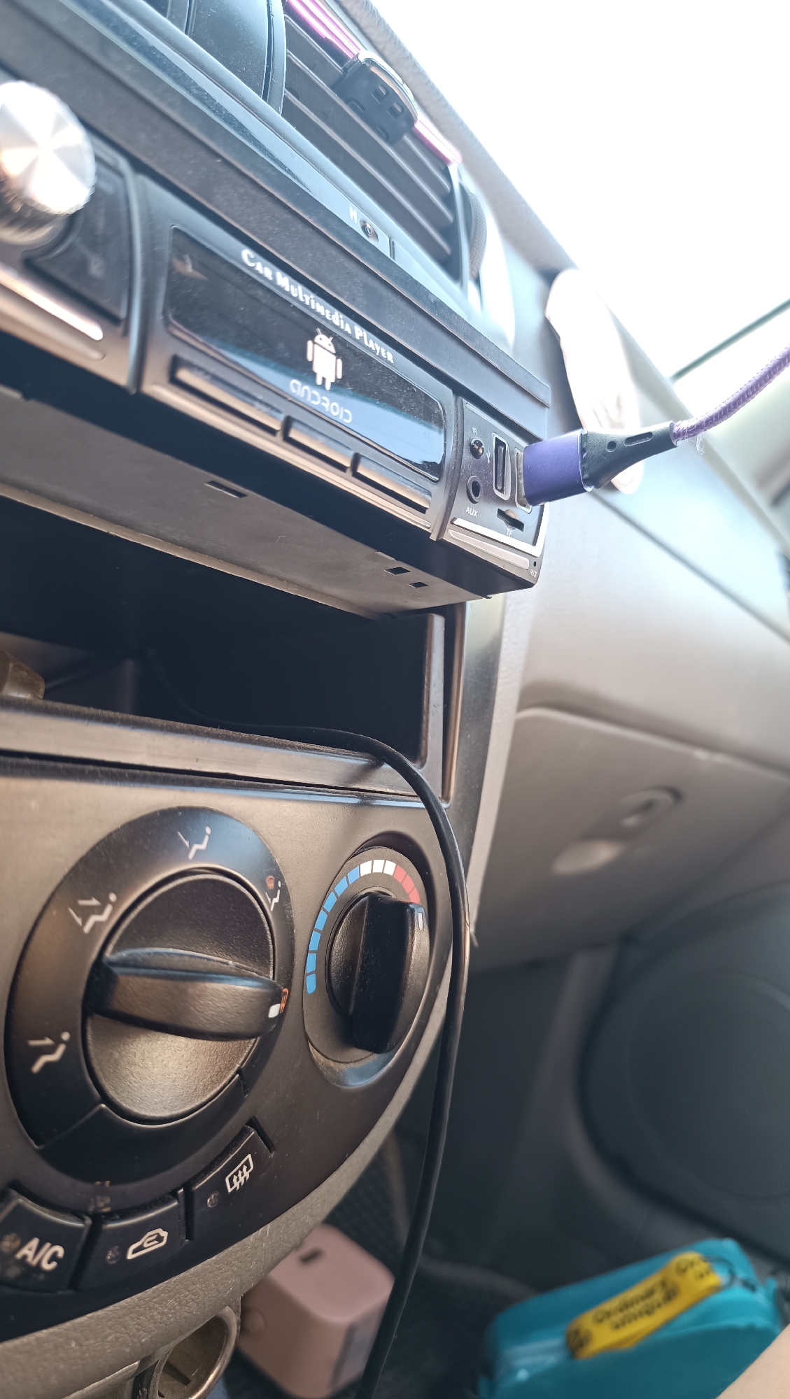 An eye-level, medium shot captures the interior of a car in Chaikovsky, Russia. The focus is on the central console, dominated by a car multimedia player with an Android logo. A purple USB cable is plugged into the player, with its other end extending out of frame. Below the player are ventilation controls, featuring two rotary knobs and several buttons. One knob controls temperature, indicated by blue and red segments, while the other likely controls fan speed or air direction. Buttons include icons for defrosting, A/C, and a circular airflow symbol. A black cable snakes down from the multimedia player towards the lower part of the console. In the background, the car's dashboard and passenger seat are visible, with a teal colored object and a pink box-like item resting on the passenger side.