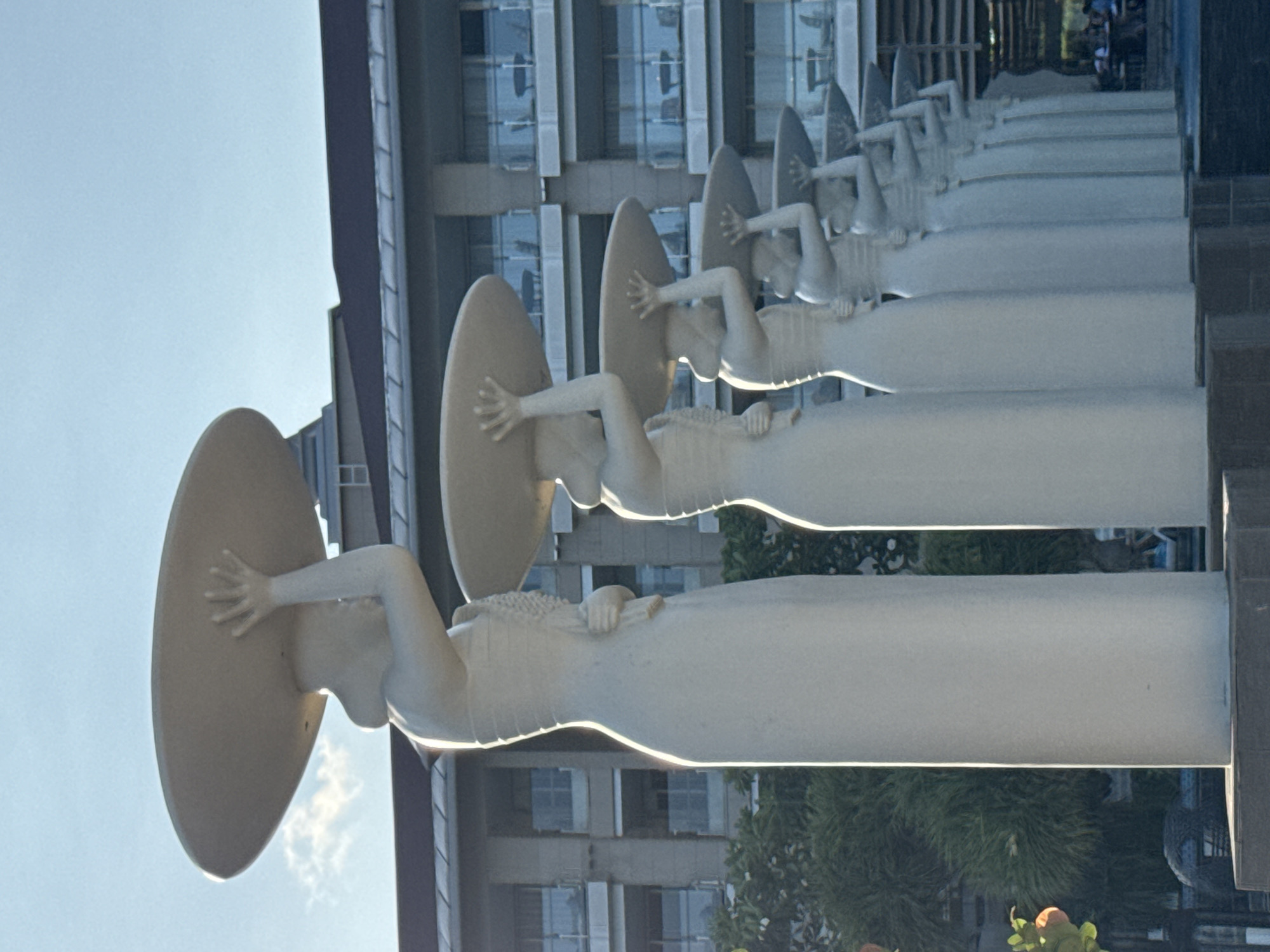 A series of identical white sculptures of reclining women are positioned in a row outdoors. Each figure has one arm raised to hold a large, flat, circular object above their head. A multi-story building with numerous windows is visible in the background. The sky is a pale blue. Based on the architectural style and vegetation, the location is likely Kuta Selatan, Indonesia.