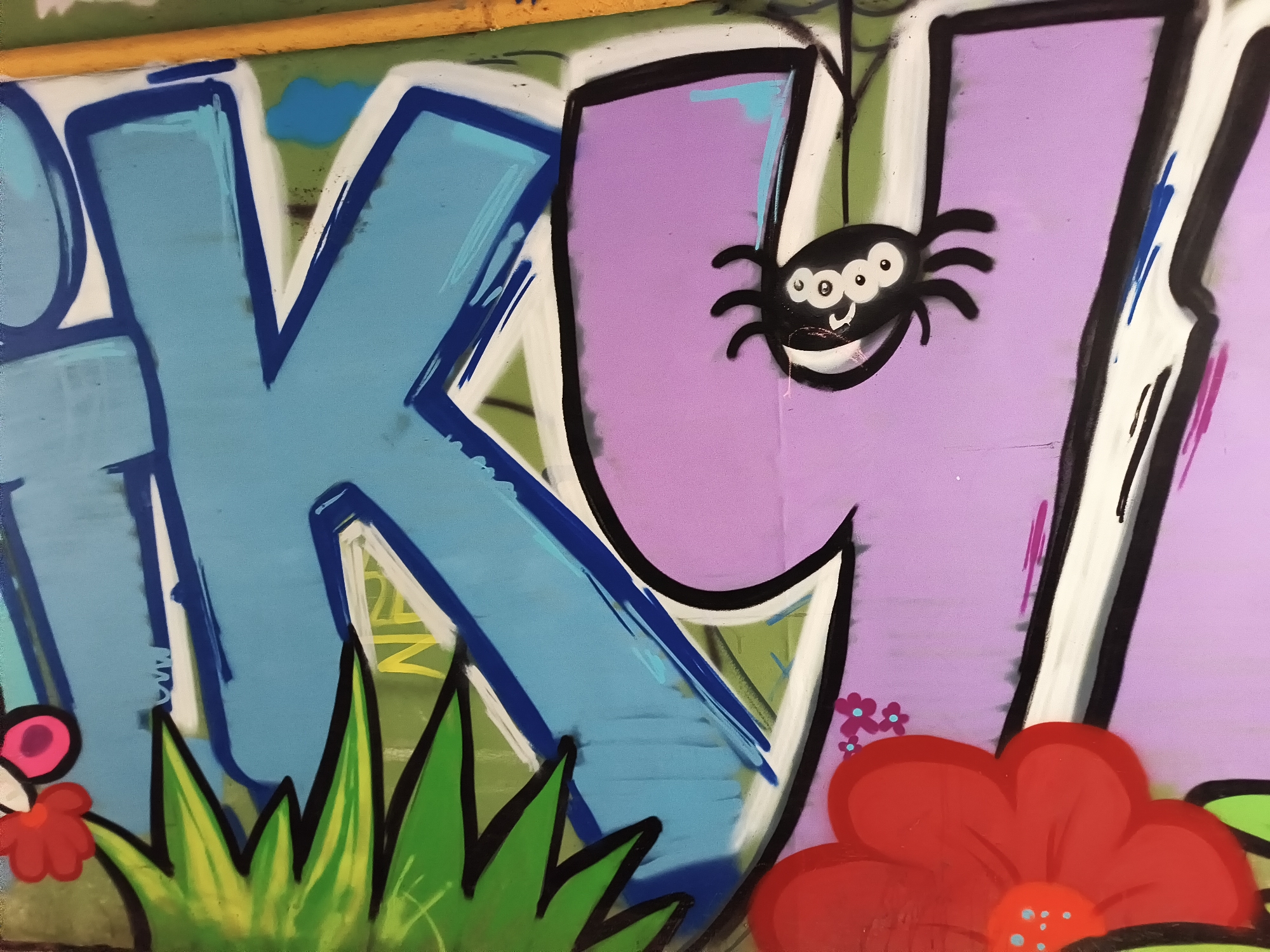 The image shows a section of graffiti art. Predominantly visible are two large block letters, "K" and "I," in light blue and lavender respectively. The letter "K" is outlined in white, with a dark blue secondary outline, and a patch of green with yellow. The letter "I" is outlined in white with a black secondary outline. A stylized cartoon spider with multiple eyes and a smile is depicted on the face of the letter "I." A red flower is present in the lower right. Some greenery is also present. The location is identified as Kyiv, Ukraine.