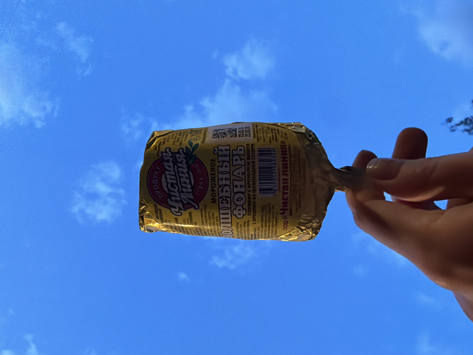 A hand is holding an ice cream bar against a backdrop of a blue sky with scattered clouds. The ice cream bar is wrapped in gold foil and bears the name "Чистая линия" (Chistaya Liniya) and the phrase "ВОЛШЕБНЫЙ ФОНАРЬ" (Volshebnyi Fonar), which means "Magical Lantern" in English. This brand and product are produced by a company called "Chistaya Liniya" (Clean Line) in Russia. The image was likely taken outdoors, in Moscow, Russia.