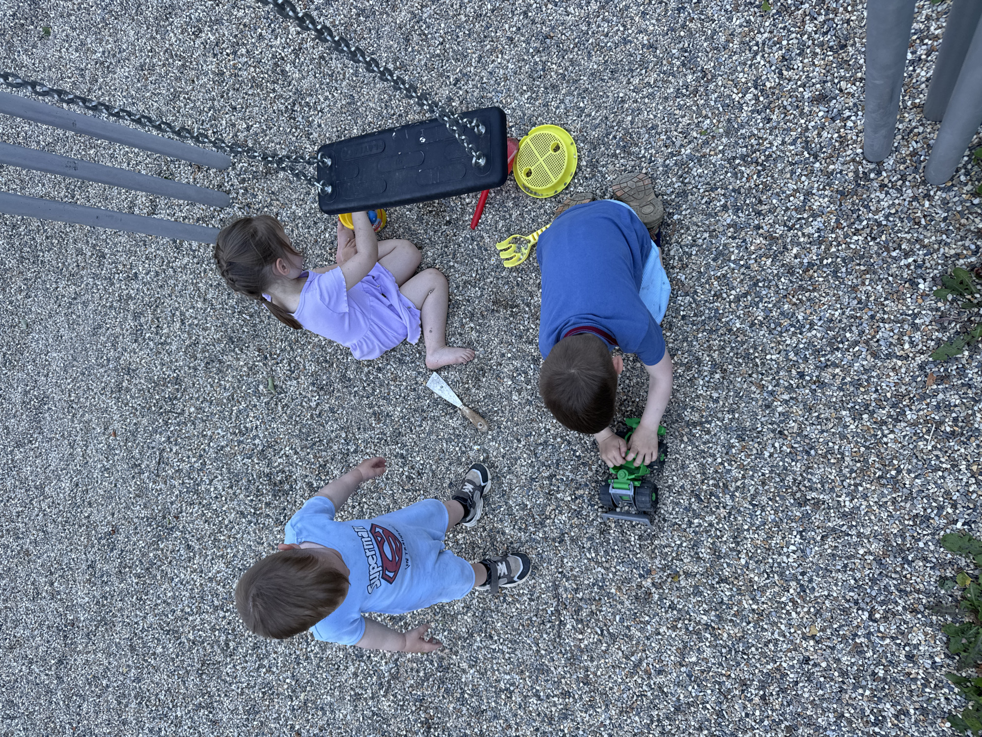 Three young children are playing on a gravel playground. A boy in a light blue Superman t-shirt and shorts stands to the left, looking down. In the center, a girl in a lavender dress is sitting near a black swing seat, holding a yellow bucket. To the right and in the foreground, a boy in a blue t-shirt and light blue shorts is playing with a green toy truck. Several other toys, including a yellow rake and a red shovel, are scattered on the gravel. Two gray metal poles are visible to the right of the swing.