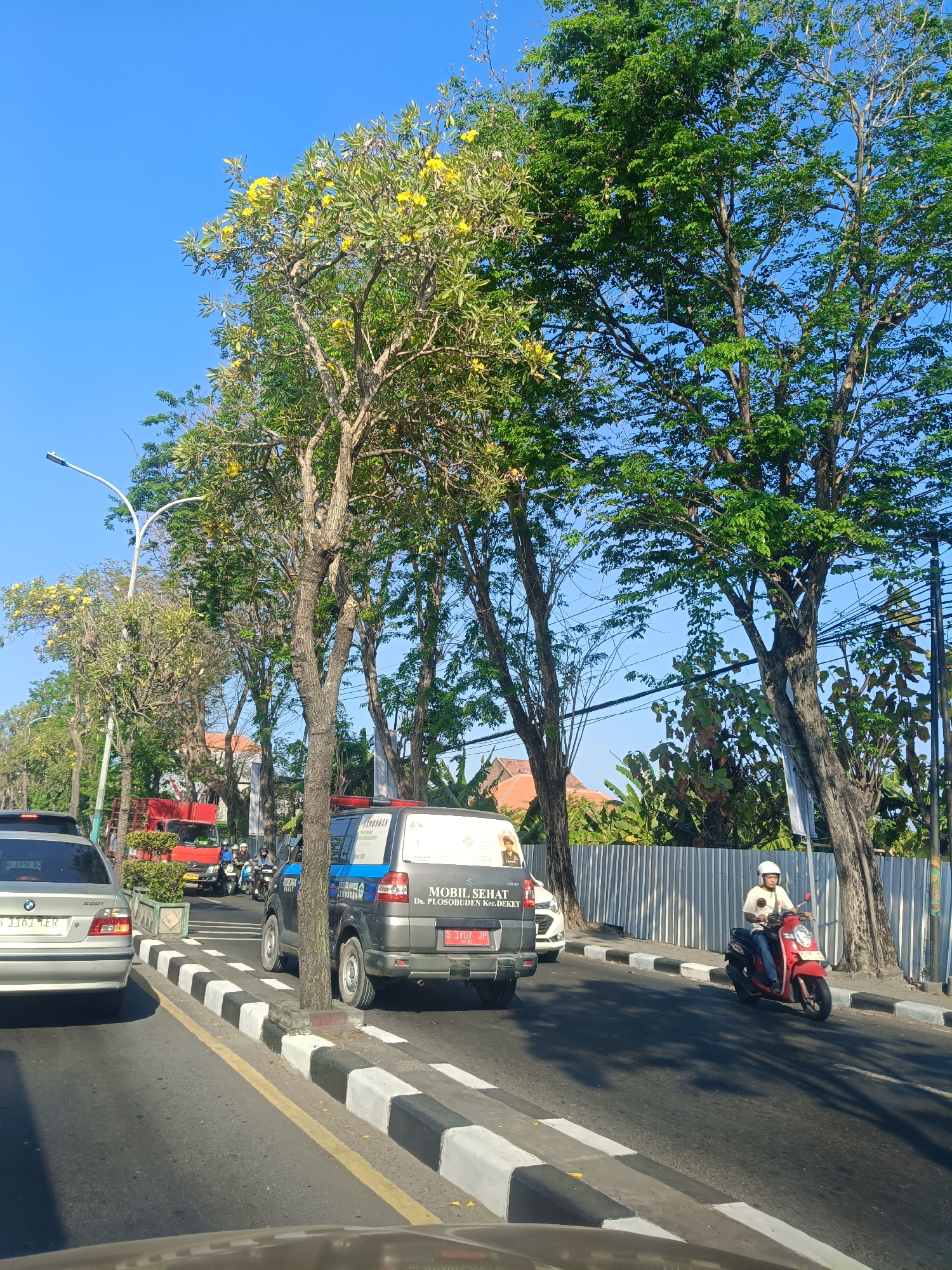 The image depicts a street scene in Klangonan, Indonesia. The sky is clear blue. On the right side of the road, a person is riding a red scooter wearing a white helmet and a light-colored shirt. The scooter is moving forward. To the left of the scooter, a black utility vehicle is parked, with text and images visible on its side. In front of the utility vehicle, two people on a motorcycle are also stopped. Further to the left, a silver sedan is in the lane of traffic. The road has a zebra crossing with black and white stripes. Trees with green foliage and some with yellow flowers line the street. A street lamp is visible on the left side of the image. In the background, there are buildings and a metal fence. The overall scene suggests a daytime street view with active traffic.
