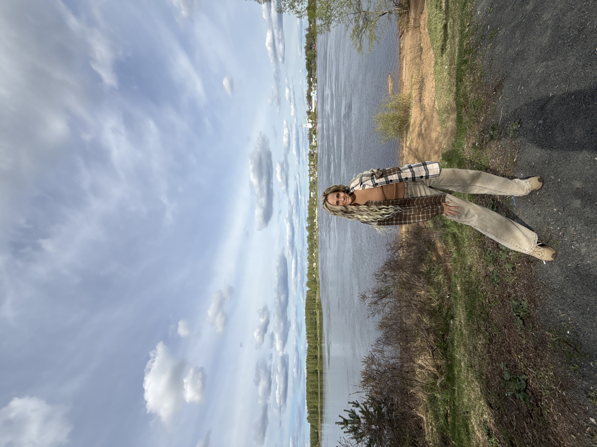A woman stands on a paved path next to a grassy area, with a large body of water behind her and a cloudy sky above. The body of water appears to be the Volga River, with the city of Заволжье, Russia, visible on the opposite bank. The woman is wearing a brown and beige plaid jacket, beige pants, and brown shoes. She has long blonde hair. The path appears to be used for walking or biking.