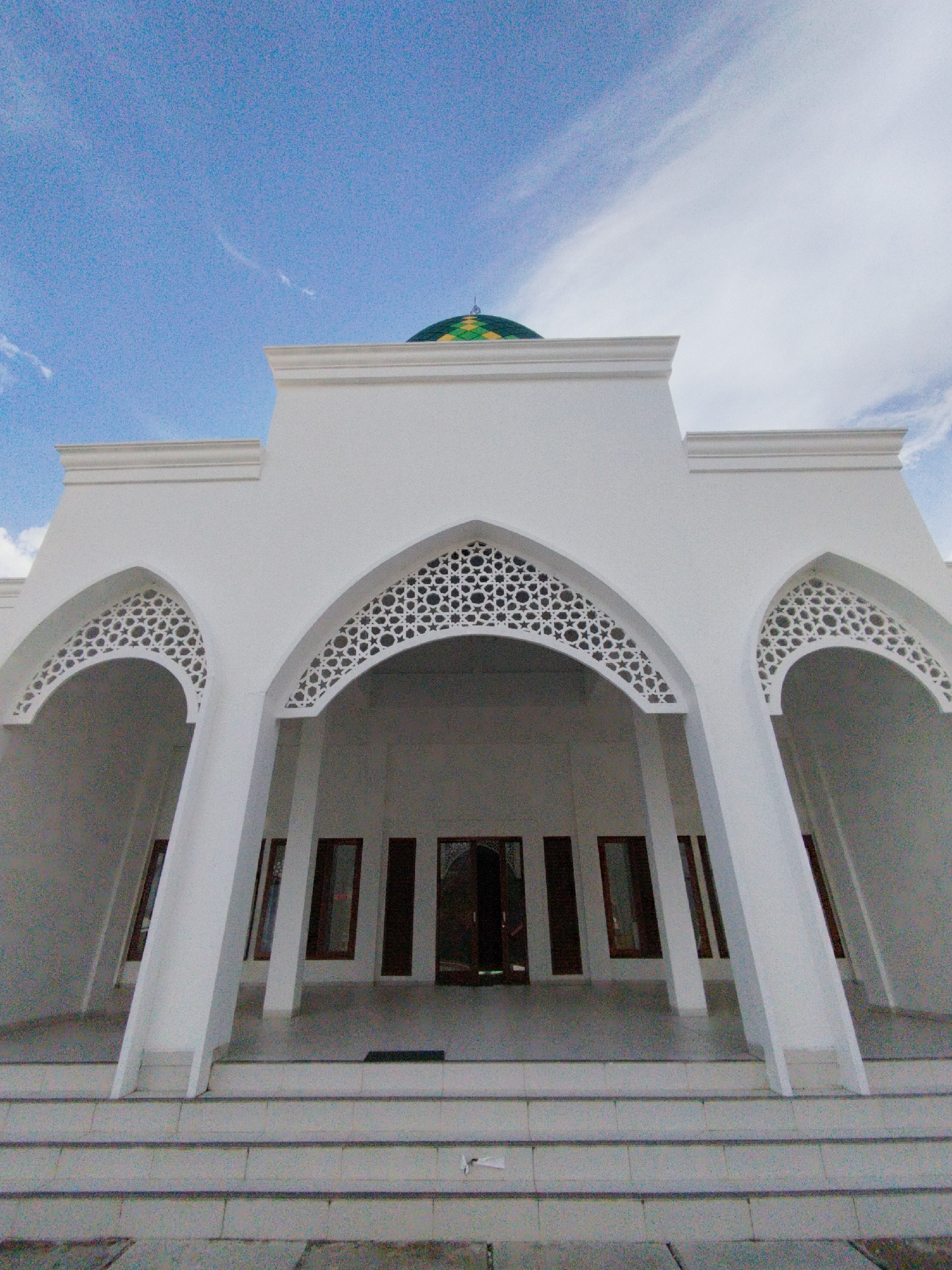 The image captures the facade of a white mosque with a green dome visible against a bright blue sky with wispy white clouds. The mosque features prominent arched entrances adorned with intricate geometric patterns. Several tall, narrow windows with dark frames are visible along the facade. A set of concrete steps leads up to the building. The foreground shows the pavement and the base of the steps, with a hint of foliage or ground cover. The location is identified as Keureuweungkrueng, Indonesia. No people are visible in the image.