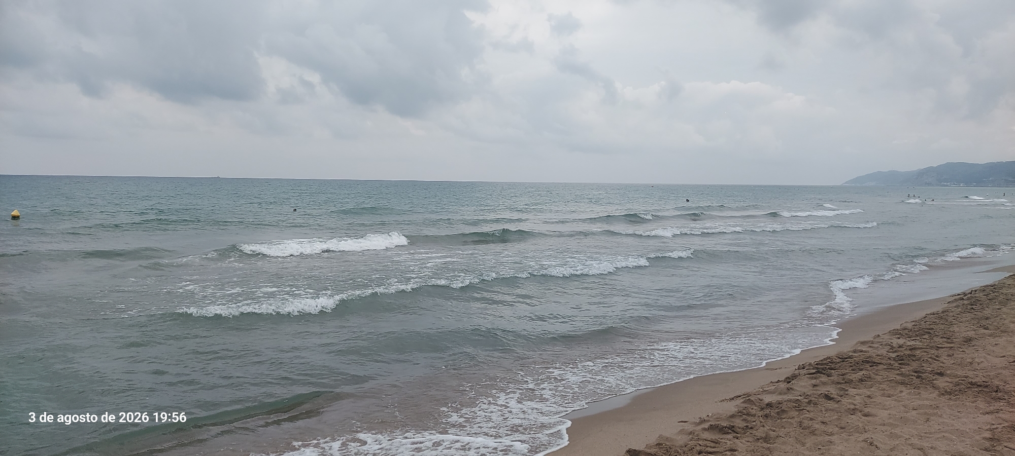 The image shows a view of the beach and sea in Castelldefels, Spain, on August 3, 2026, at 19:56. The sky is overcast with gray clouds. The sea is a muted blue-green, with several sets of waves breaking on the shore. The beach in the foreground is sandy, with some textured patterns from the sand. There are a few small figures visible in the water, appearing to be people swimming or wading, and a yellow buoy is visible in the distance to the left. The coastline extends into the distance to the right, with a faint outline of hills or mountains under the cloudy sky. The timestamp "3 de agosto de 2026 19:56" is visible in the lower left corner.