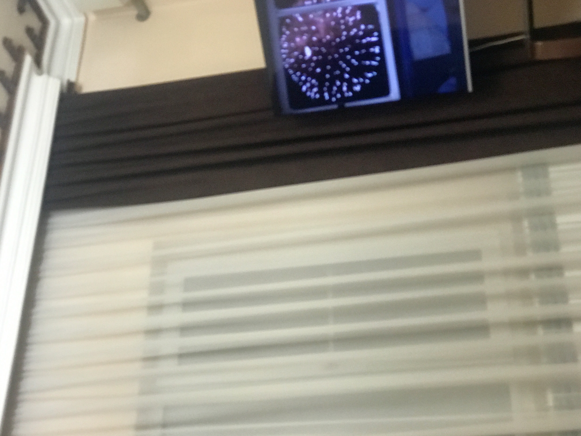 A blurred image shows a television screen displaying an image resembling fireworks above closed, horizontal blinds. The location is unknown and cannot be determined from the image. No people, actions, or interactions are visible.