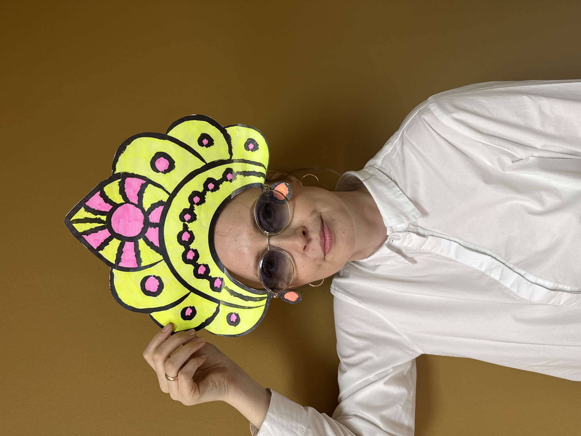 A woman is holding a yellow and pink paper flower in front of a brown background. She is wearing a white button-down shirt and sunglasses. Her hand is visible, with a ring on her finger. The location is not discernable from the image.