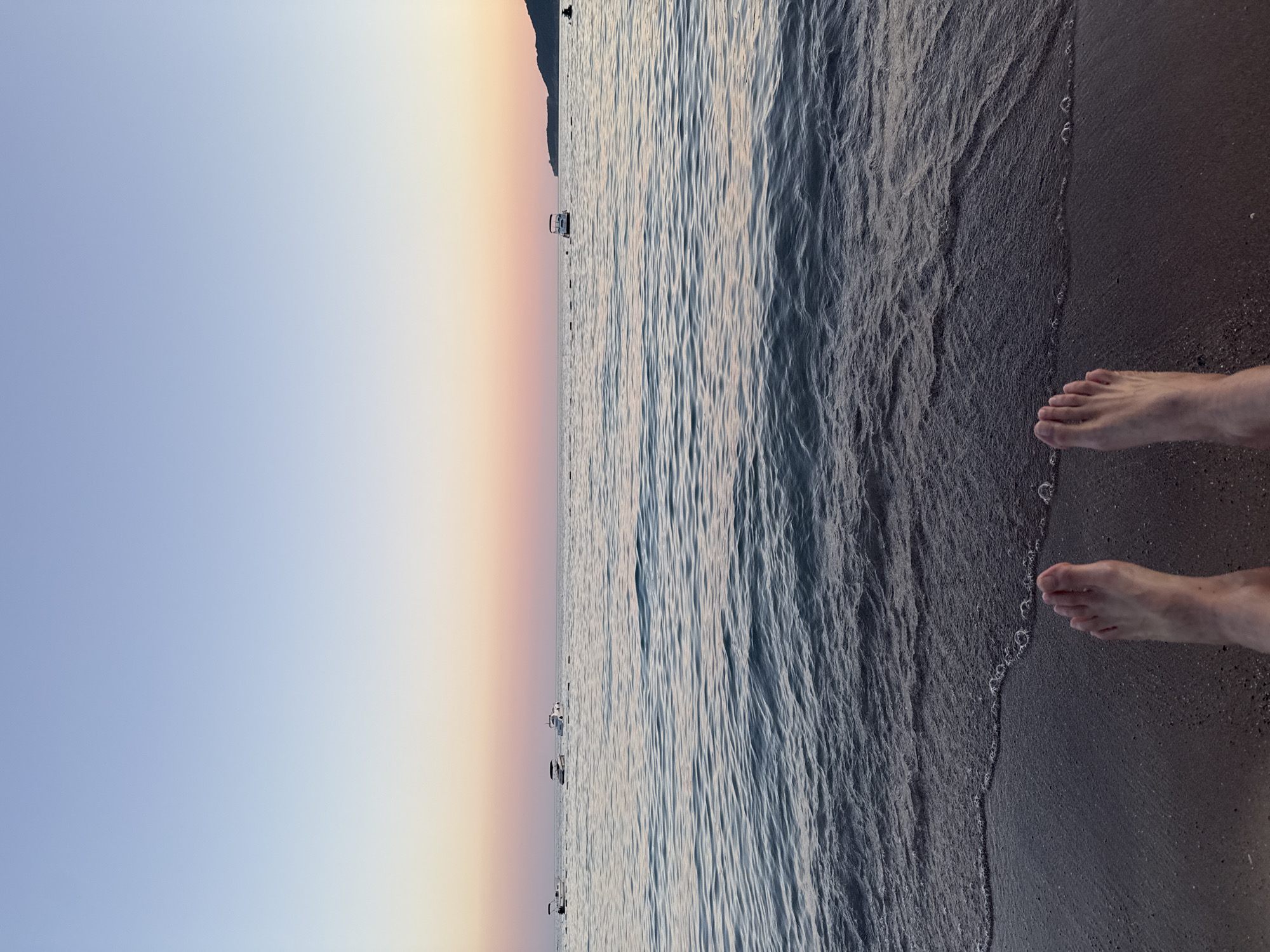 The image depicts a serene beach scene at sunset in Rafailovići, Montenegro. The foreground shows the bare feet of an individual standing on the wet sand at the edge of the water. Gentle waves are lapping at the shore, creating a thin line of foam and bubbles. The sea extends to the horizon, where it meets a sky painted with soft hues of blue, pink, and orange, characteristic of twilight. Several small boats are anchored in the calm water, adding a touch of activity to the tranquil setting. In the distance, a hilly coastline is visible, silhouetted against the colorful sky. The overall atmosphere is peaceful and relaxing, suggesting a moment of quiet contemplation by the sea.