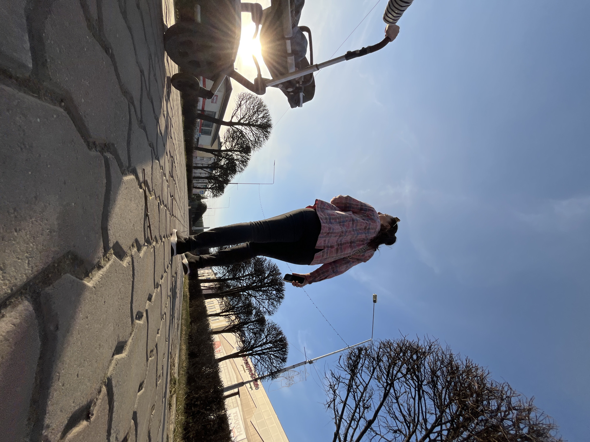 The image captures an outdoor scene from a very low angle, looking upwards from a paved pathway in Zelenogorsk, Russia, on a bright, sunny day.
A person, likely female, is featured prominently, standing on the grey interlocking block pavement. She is dressed in a plaid shirt (featuring pink/red and light-colored squares), dark pants, and dark sneakers with white soles. She holds a dark smartphone in her right hand.
In the immediate foreground, a baby stroller is partially visible, with the sun positioned directly behind it, causing a prominent lens flare. The stroller's handle, equipped with a striped grip, is visible.
The background features a clear, vivid blue sky. Bare-branched trees, suggesting late winter or early spring, line the pathway. A street lamp post stands tall. In the distance, several buildings are visible, including one with a reddish roof and another light-colored commercial structure. Faint text, possibly "TEXN", can be seen on one of the buildings.
The bright sunlight and clear sky indicate it is daytime, likely mid-morning or afternoon. The scene depicts a casual outdoor stroll, with the low camera perspective offering an expansive view of the sky and surroundings.