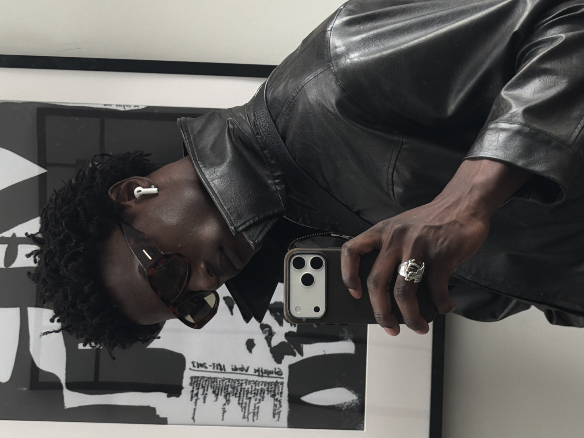 A dark-skinned man is taking a selfie. He is wearing a black leather jacket, sunglasses, and an earbud in his left ear. He is also wearing a large, ornate ring on his right hand. The phone is dark with three cameras on the back. He is holding the phone up to a mirror, which reflects a black and white abstract picture. The location is likely indoors and cannot be identified as being in London.