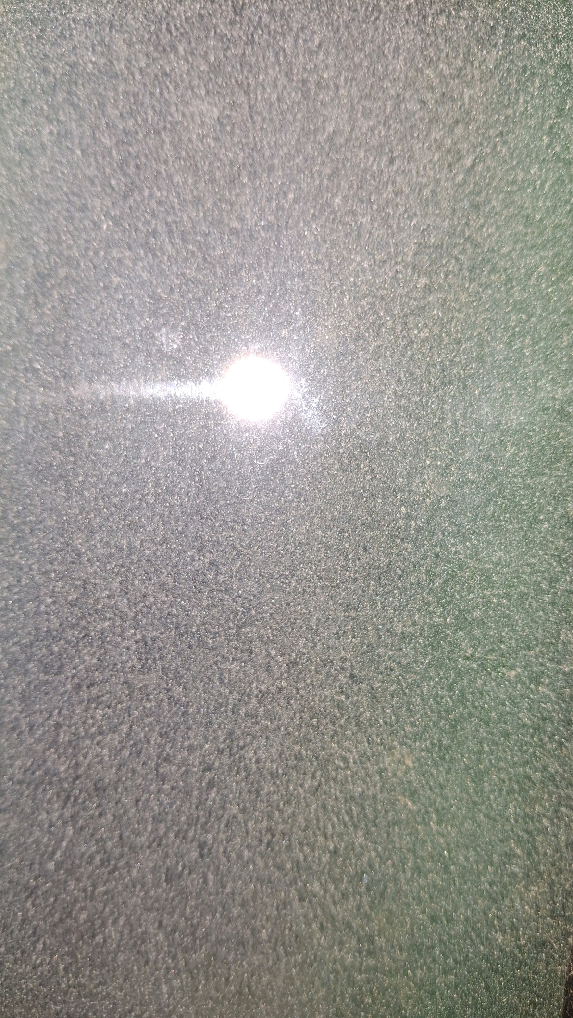 The image shows a close-up, macro view of a textured surface, possibly glass or a similarly reflective material, with a bright light source creating a glare. The surface appears to be speckled or granular. The lighting suggests it could be illuminated by a flash or a bright point of light. The colors are muted, with a dominant grayish tone and a hint of green in the upper right portion of the frame. There are no discernible people, objects, or actions within the image. The location in Sumberrejo, Indonesia, is not visually represented in this abstract photograph.