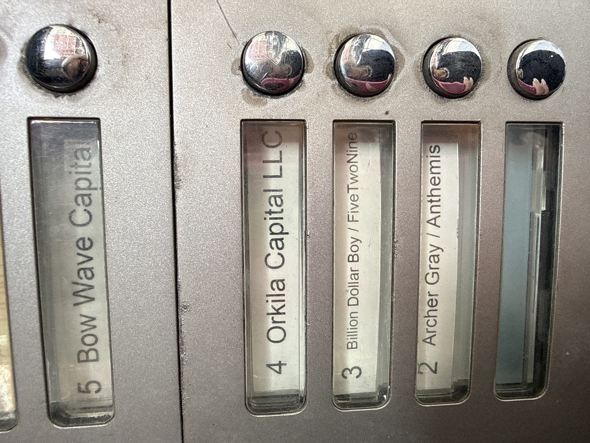 The image shows a control panel with labels for different funds or entities. From left to right, the labels are: "5 Bow Wave Capita," "4 Orkila Capital LLC," "3 Billion Dollar Boy / FiveTwoNine," and "2 Archer Gray / Anthemis." There is an unlabeled slot on the far right. The panel appears to be made of metal, with buttons above each label. The buttons are round and reflective. The labels are printed in a simple sans-serif font. There is no explicit information about the location in New York City.