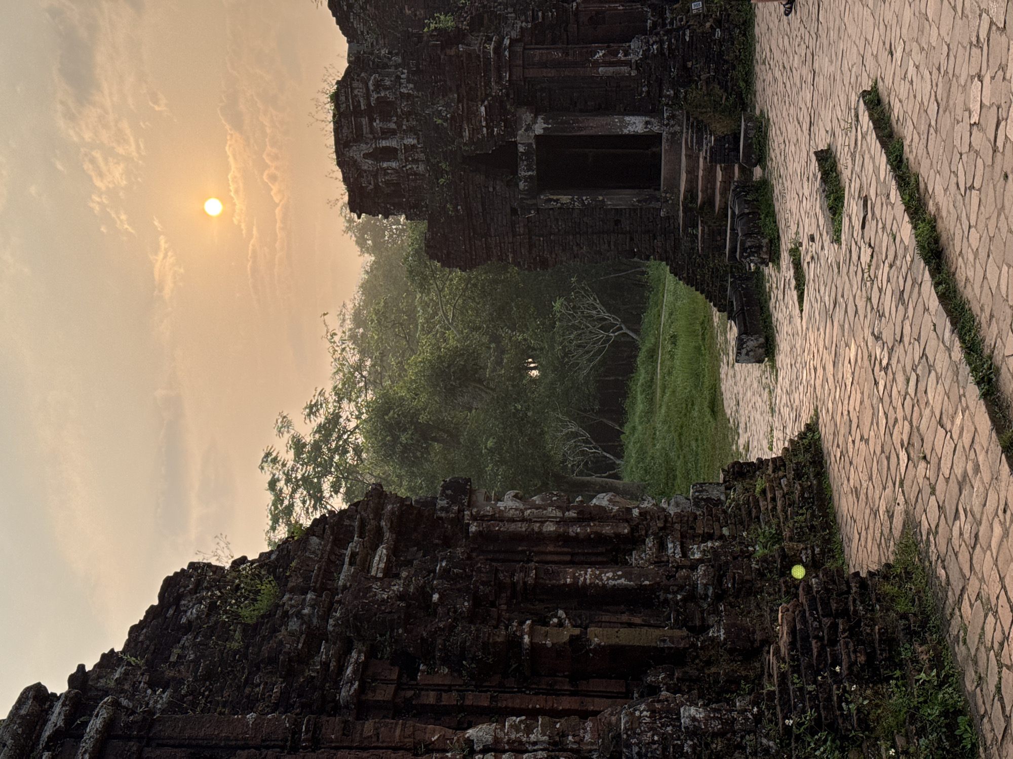 The image shows a pathway framed by ancient brick structures. The pathway leads to a grassy area with trees. The sun is visible through the hazy sky. The scene is likely located in My Son Sanctuary in Đà Nẵng, Vietnam, a UNESCO World Heritage site comprising Hindu temple ruins dating back to the Champa kingdom. A cobblestone path runs alongside ruins. A golf ball is visible on the right side of the ruins.