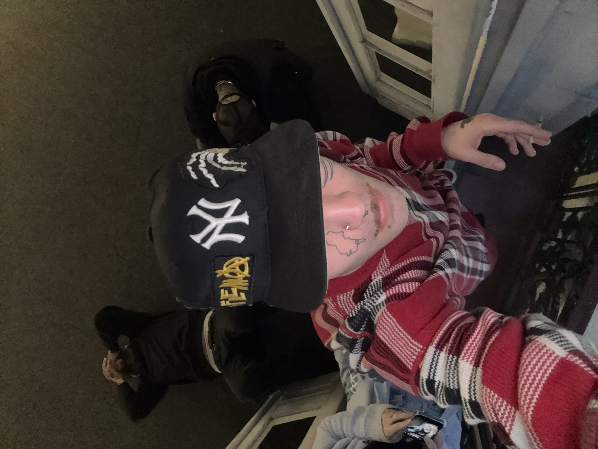 Three people are present. One person, presumed to be taking the photograph, is wearing a black baseball cap with a New York Yankees logo and a gold "FEMO" patch, a red and gray plaid shirt, and has facial piercings and tattoos. This person is holding a phone or small electronic device.
Two other people are visible lying on the ground. One is wearing dark clothing and covering their face with their hands, while the other is wearing black clothing and a face covering.
The scene appears to be outdoors on a balcony or raised platform, with a building and a street below. The location is Buenos Aires, Argentina; however, there is no supporting evidence within the image to corroborate this claim.