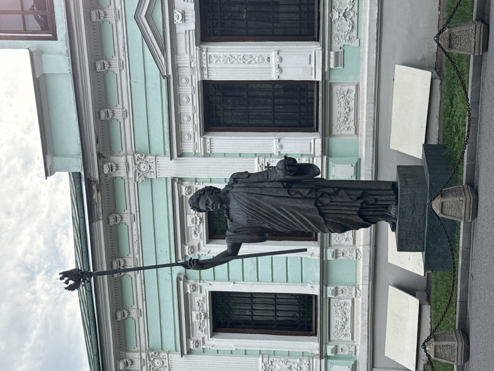 The image shows a bronze statue of a woman holding a scepter and an orb in front of a light green building with white ornamentation. The statue is positioned on a dark pedestal. The building has multiple windows with dark frames and bars, and decorative moldings around the windows and roofline. In the foreground, there is a grey paved area with a curb, a patch of green grass, and decorative stone bollards connected by chains. The sky is overcast.
This statue is believed to be of Catherine the Great, located in Moscow, Russia. It is positioned in front of the Catherine Palace (Ekaterininskiy dvorets), which is a building within the Kremlin complex in Moscow.