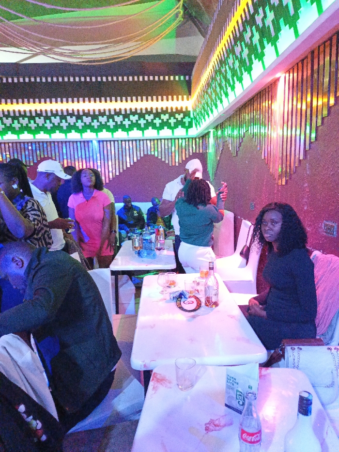 A group of people gathers at a bar with tables and chairs. Some are standing and talking, while others are seated. Drinks and bottles are visible on the tables. The bar has a modern design with decorative lighting. The person sitting on the right side of the image is wearing a dark dress, looking toward the camera. The atmosphere is lively and social.