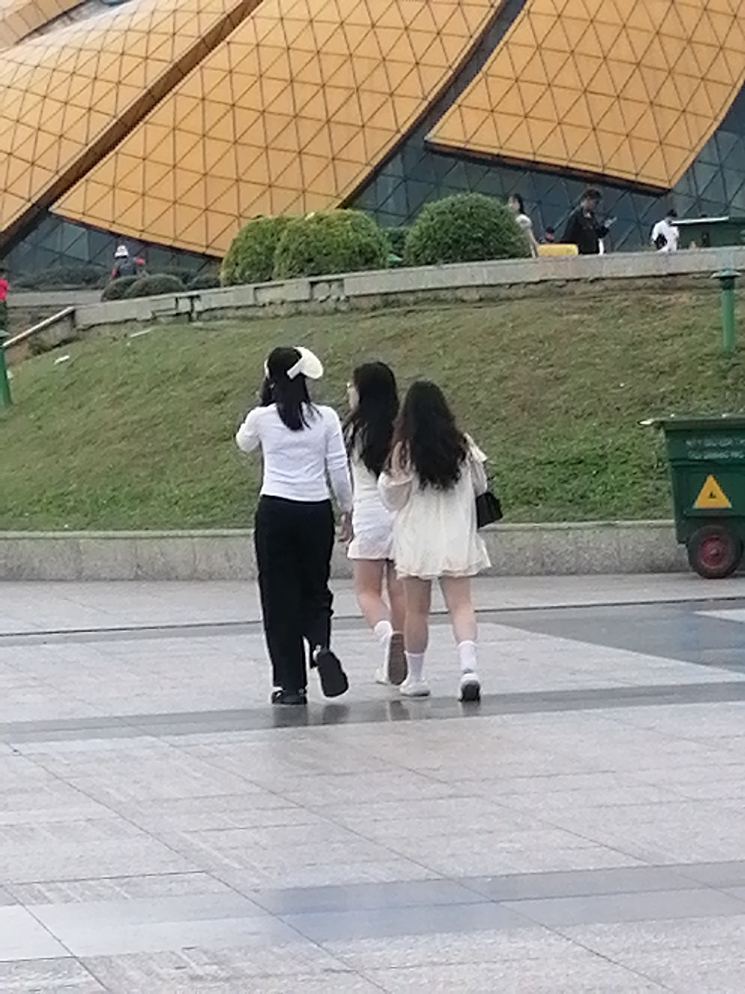 The image captures a view of the Quang Trung street area in Da Lat, Vietnam, with a distinctive architectural structure featuring a yellow, triangular-patterned facade dominating the background. In the mid-ground, three young women are seen walking away from the camera on a paved walkway. The woman on the left wears a white long-sleeved shirt and black pants, accessorized with a white bow on her head. The middle woman is dressed in a white dress, and the woman on the right also wears a white dress. They are walking past a grassy embankment. To the far right of the image, a green trash bin with a yellow triangular symbol is visible. The foreground consists of a paved plaza.