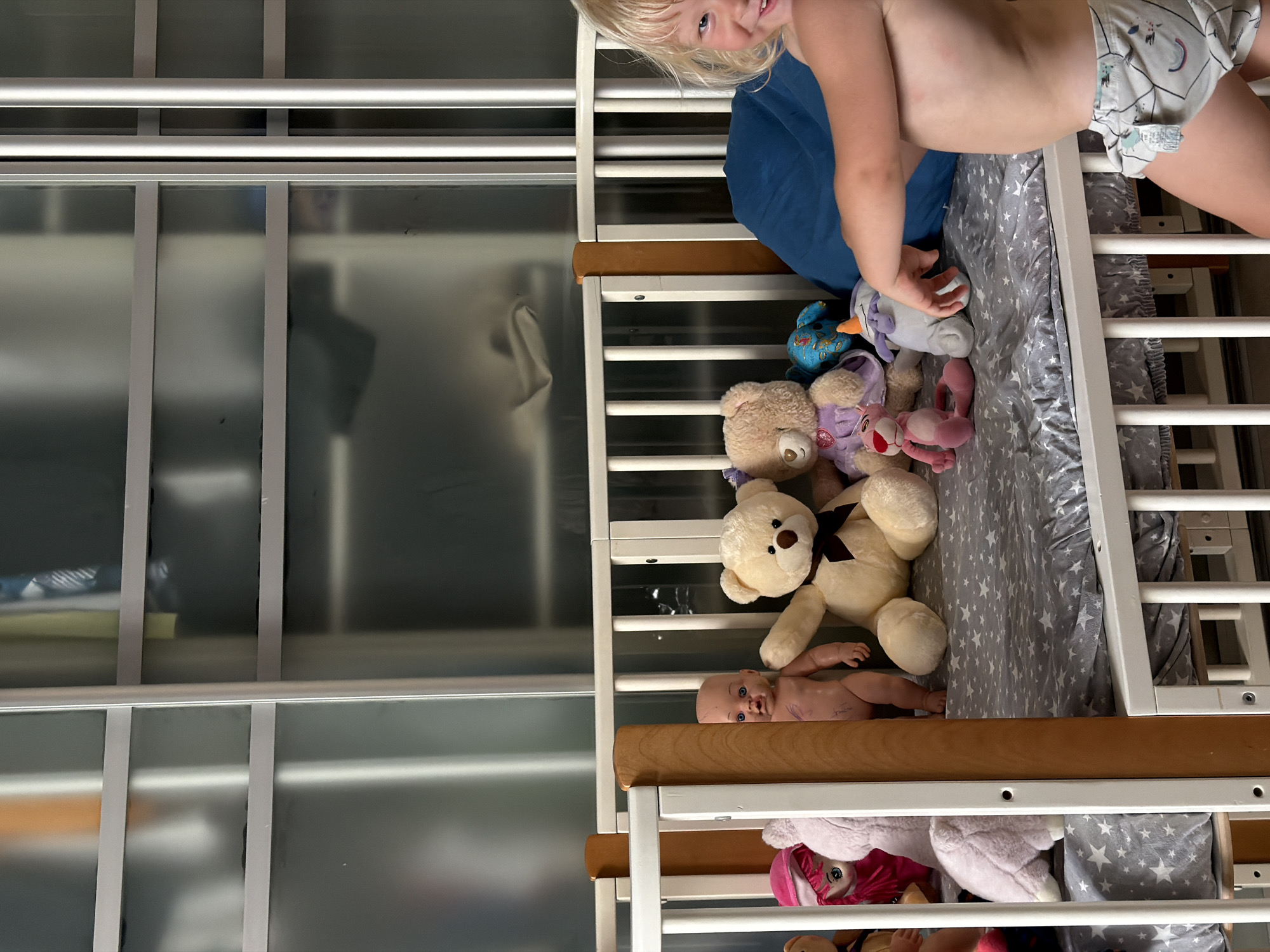 The image captures a top-down view of a child's crib filled with stuffed animals and a doll, with a toddler peeking into the frame from the top. The toddler, with blonde hair, is smiling broadly at the camera. They are wearing a patterned diaper and are partially visible from the chest up. The crib is white with wooden trim, and the bedding is a gray fabric with white stars.
Several stuffed animals are scattered around the crib, including a large tan teddy bear with a dark bow, a smaller tan teddy bear, a purple stuffed animal that appears to be a unicorn or similar mythical creature, and a pink stuffed animal with large eyes. A baby doll with blue eyes is lying on its back in the crib, positioned between the teddy bears. At the bottom of the crib, more stuffed animals are visible, including a pink stuffed animal wearing a hooded garment.
The setting appears to be indoors, likely a child's bedroom or nursery, given the presence of the crib and toys. The background to the left of the crib shows a reflective surface with vertical lines, possibly a closet or wardrobe with mirrored doors. The lighting suggests it might be daytime or artificial lighting is used. The overall mood is playful and cheerful, primarily conveyed by the toddler's expression. No visible text is present in the image. The provided location context is Sochi, Russia.