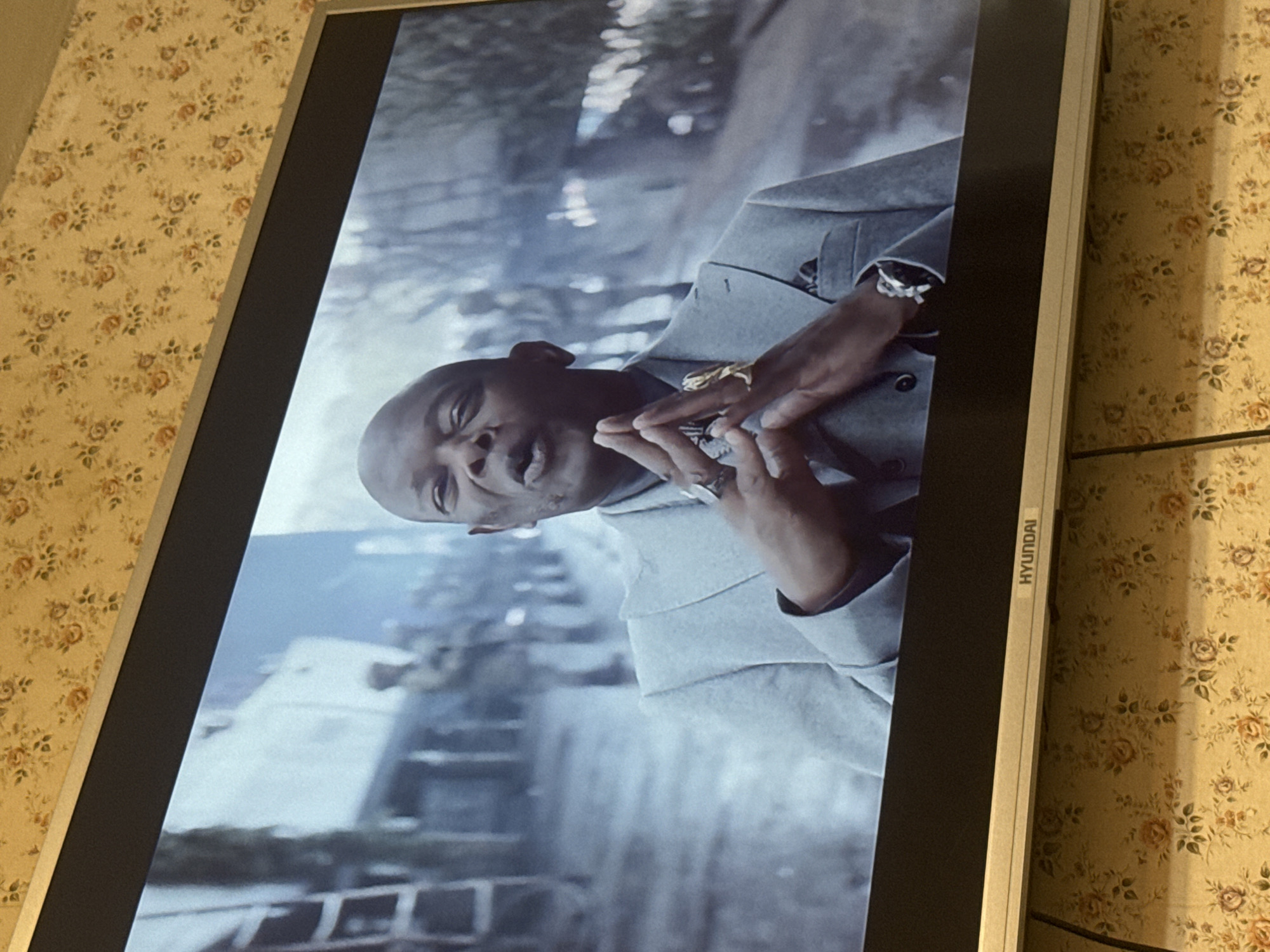A television displays a medium shot of a Black man wearing a light-colored suit and adorned with jewelry, including rings and a watch. The background appears to be an urban setting. The television, branded "HYUNDAI" along the edge, is set against a backdrop of floral wallpaper. The location in the city of Mytishchi, Russia is not provided.