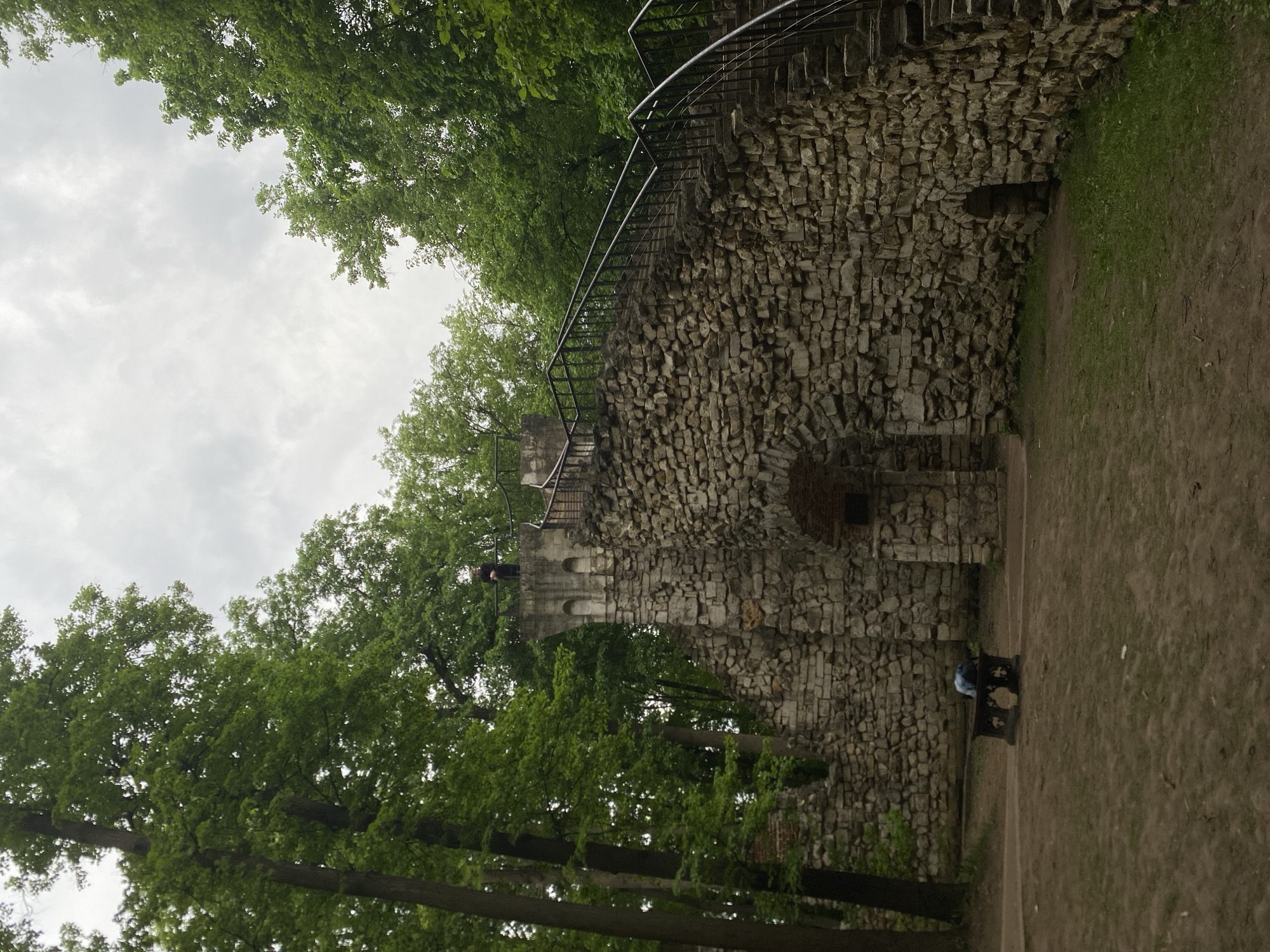 The image shows a stone wall with a metal railing and stairs leading up to a tower. The wall is made of irregular stones and has several arched openings. There is a person standing on the tower and another person sitting on a bench near the base of the wall. The wall is surrounded by trees, and the sky is cloudy. The location appears to be a park or garden in an urban area.