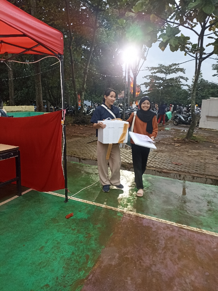 Two young women stand on a wet, concrete surface. The woman on the left is holding a white box with gold lettering. She is wearing a navy blue collared shirt, tan pants, and black shoes. The woman on the right is holding a white folder with a pink background. She is wearing a brown sweater over a white headscarf and black pants. The woman is wearing beige sandals. They are standing in front of a red canopy with a black metal frame. There are trees in the background and other people in the distance. The image was taken at the location of coordinates -6.931, 107.776, which is located in the city of Jatinangor, Indonesia.