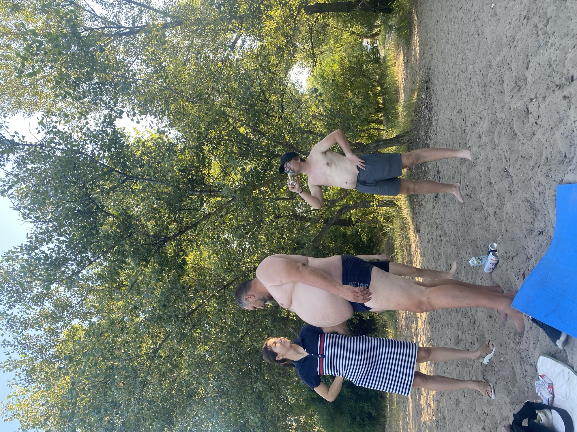 The image depicts an outdoor scene on a sandy area with trees in the background. Three individuals are visible. On the left, a woman wearing a striped dress and sandals stands with her hand on her hip, looking towards a man. The man, positioned in the center, is shirtless and wearing black swimming trunks. He has a large abdomen and is looking at the woman. To the right, a man wearing glasses and grey shorts is drinking from a can. He is shirtless and has his hand on his hip. Several cans and cigarette packs are scattered on the sand in the foreground. The environment appears to be a park or recreational area with lush green trees and a clear sky. Based on the visual context, the location is likely a beach along the Dnipro River in Dnipro, Ukraine.