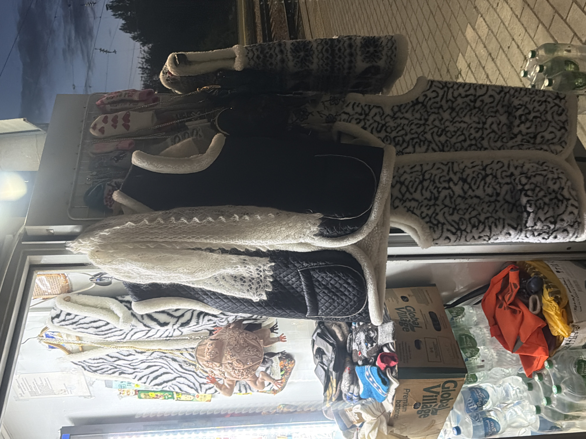 This is a photograph of a small kiosk or street stall in Balashov, Russia, at dusk. The stall displays various clothing items, including several sleeveless vests with white trim and patterned fabrics. A white, lacy scarf is draped over one of the vests. To the left, a zebra-striped vest and a unique, frog-shaped purse are hanging. Below the clothing, stacks of bottled water are visible, along with a cardboard box labeled "Global Village." In the background, there are train tracks and trees, and the sky is a dusky blue with visible power lines. The ground is paved with interlocking bricks. There are no people visible in the frame.