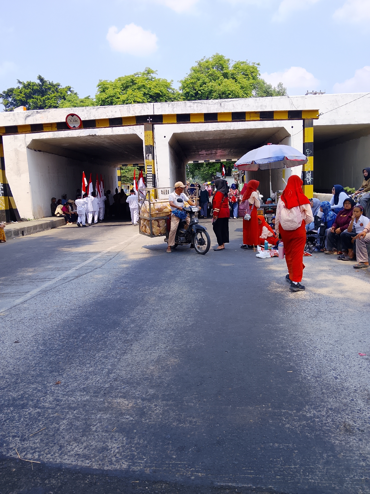 This image depicts a street scene under an overpass in Kertosono, Indonesia, likely during a daytime event. A group of people, some wearing white uniforms and carrying Indonesian flags, are gathered under the overpass. To the right, several individuals are seated, some under a blue and white umbrella, suggesting a gathering or observation point. A man on a motorcycle, carrying a large basket of what appears to be bread or pastries, is in the foreground. The overpass has warning signs indicating a height limit of 3.3 meters. The road surface is asphalt, with markings visible. The environment appears to be urban with trees visible in the background.